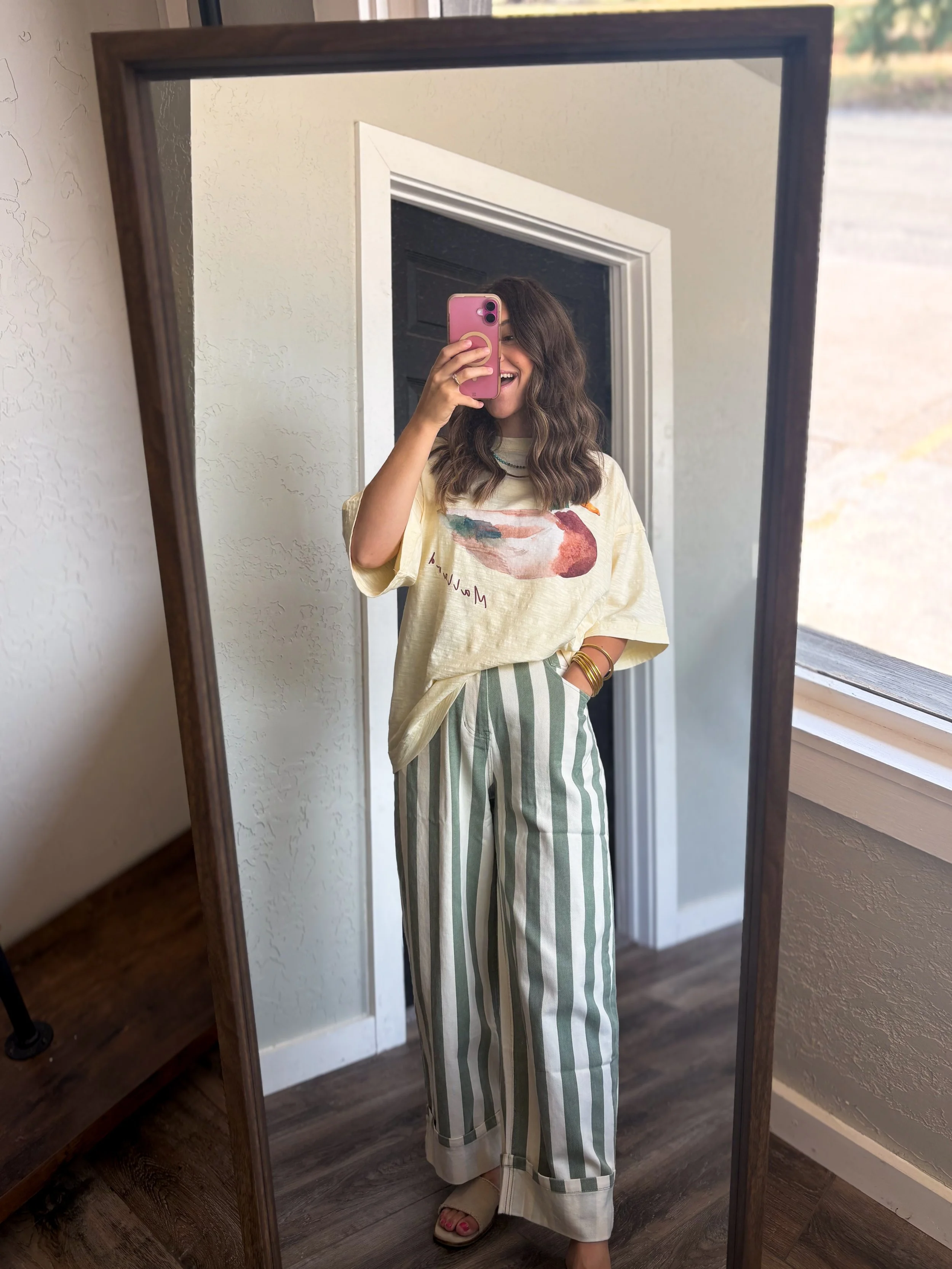 A woman smiling taking a selfie in a full-length mirror. She is wearing beige wedge sandals, light-colored top with a colorful design, and striped pants.