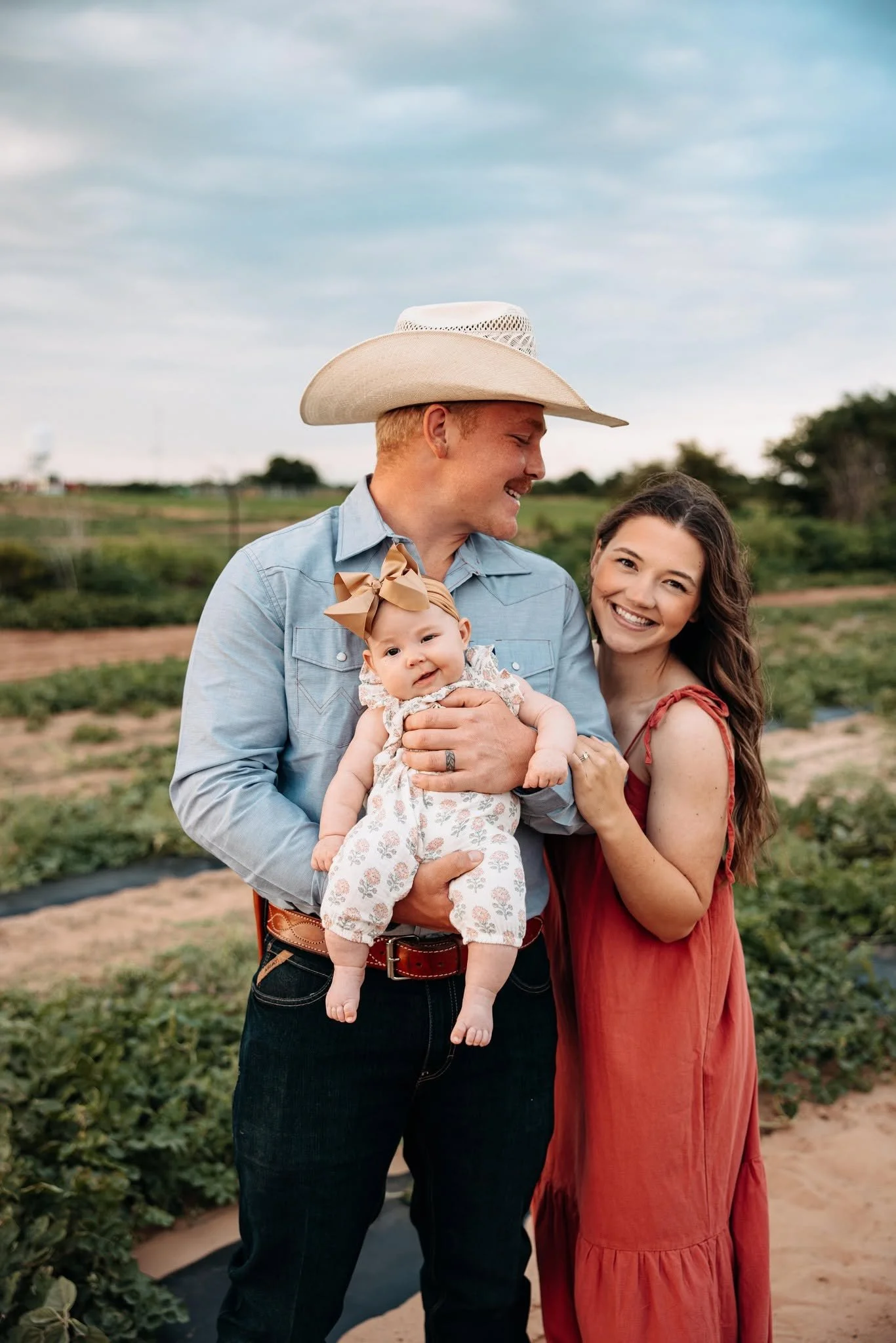 A smiling family of three, including a man wearing a cowboy hat, a woman in a red dress, and a baby with a large bow, standing outdoors in a farm or garden setting with green plants and a cloudy sky.