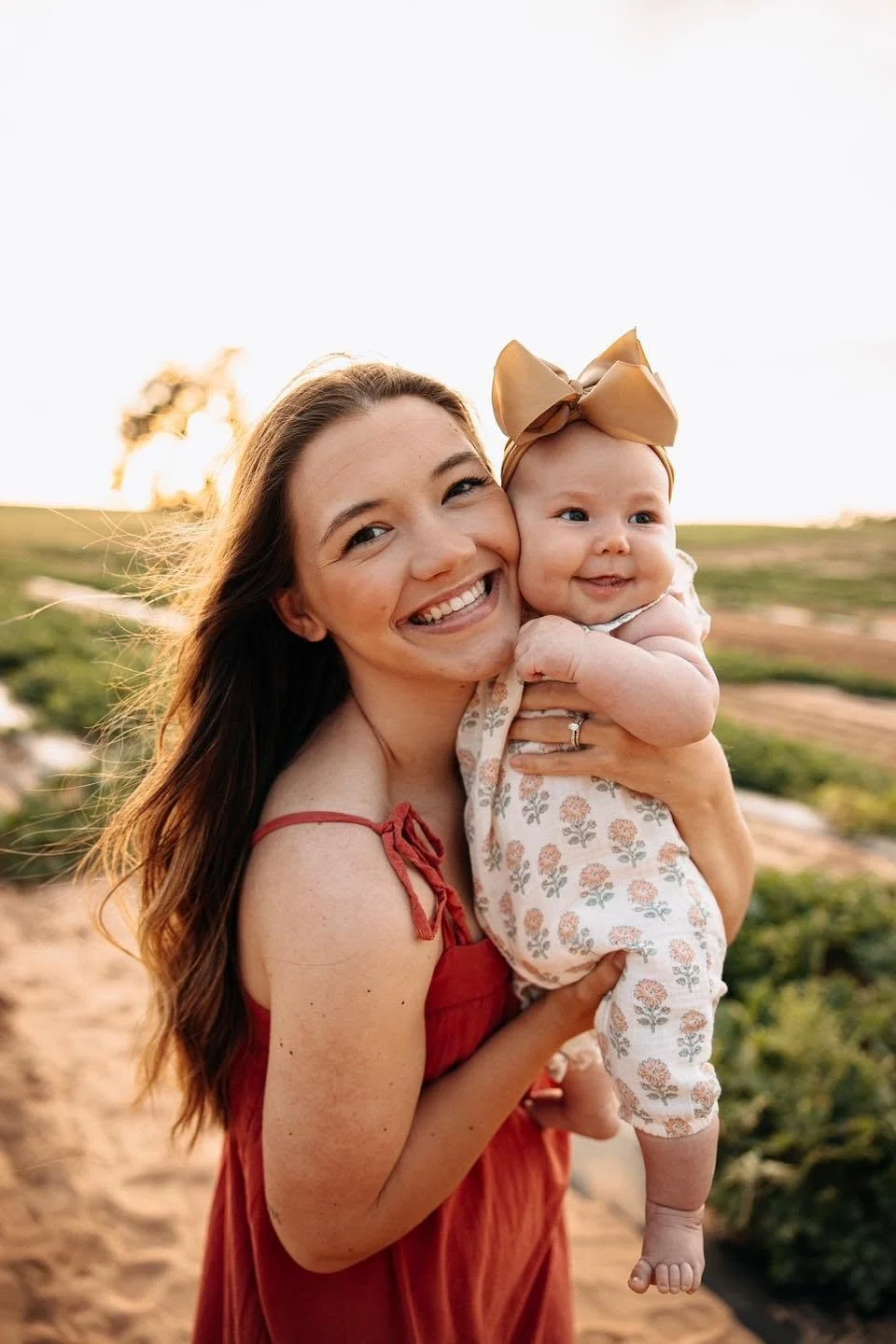 Smiling woman holding a baby girl outdoors during sunset, field background.