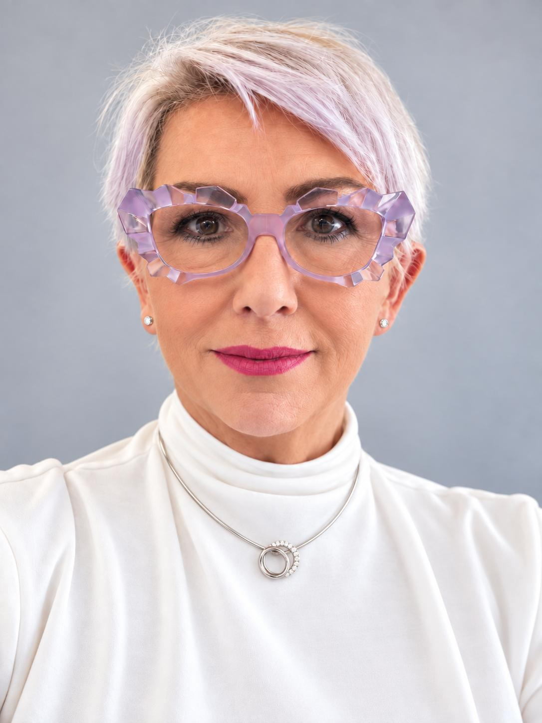 Portrait of an older woman with short lavender hair, geometric pink glasses, white blouse, and silver necklace