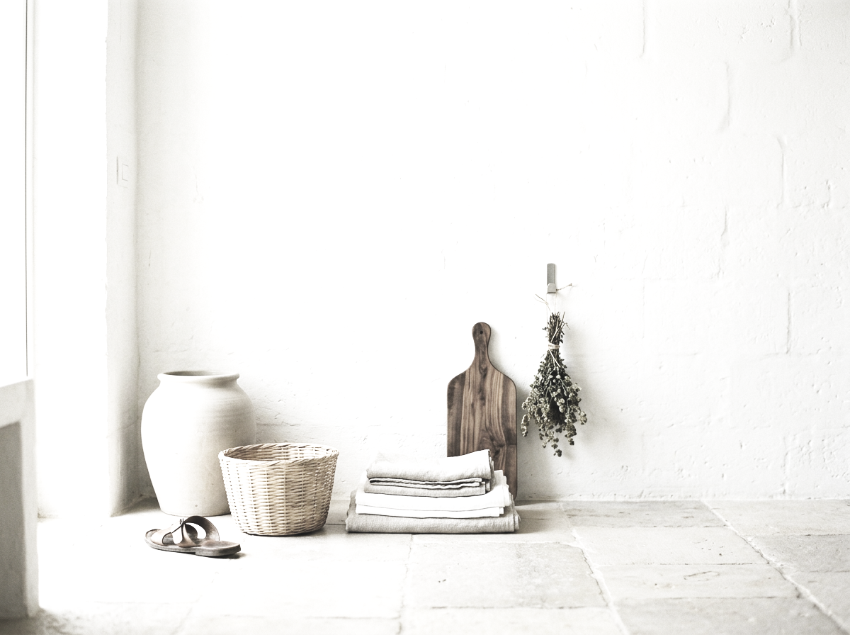 Sunlit whitewashed room with a clay vase, wicker basket of folded linens, leather sandals and dried herbs on a stone floor