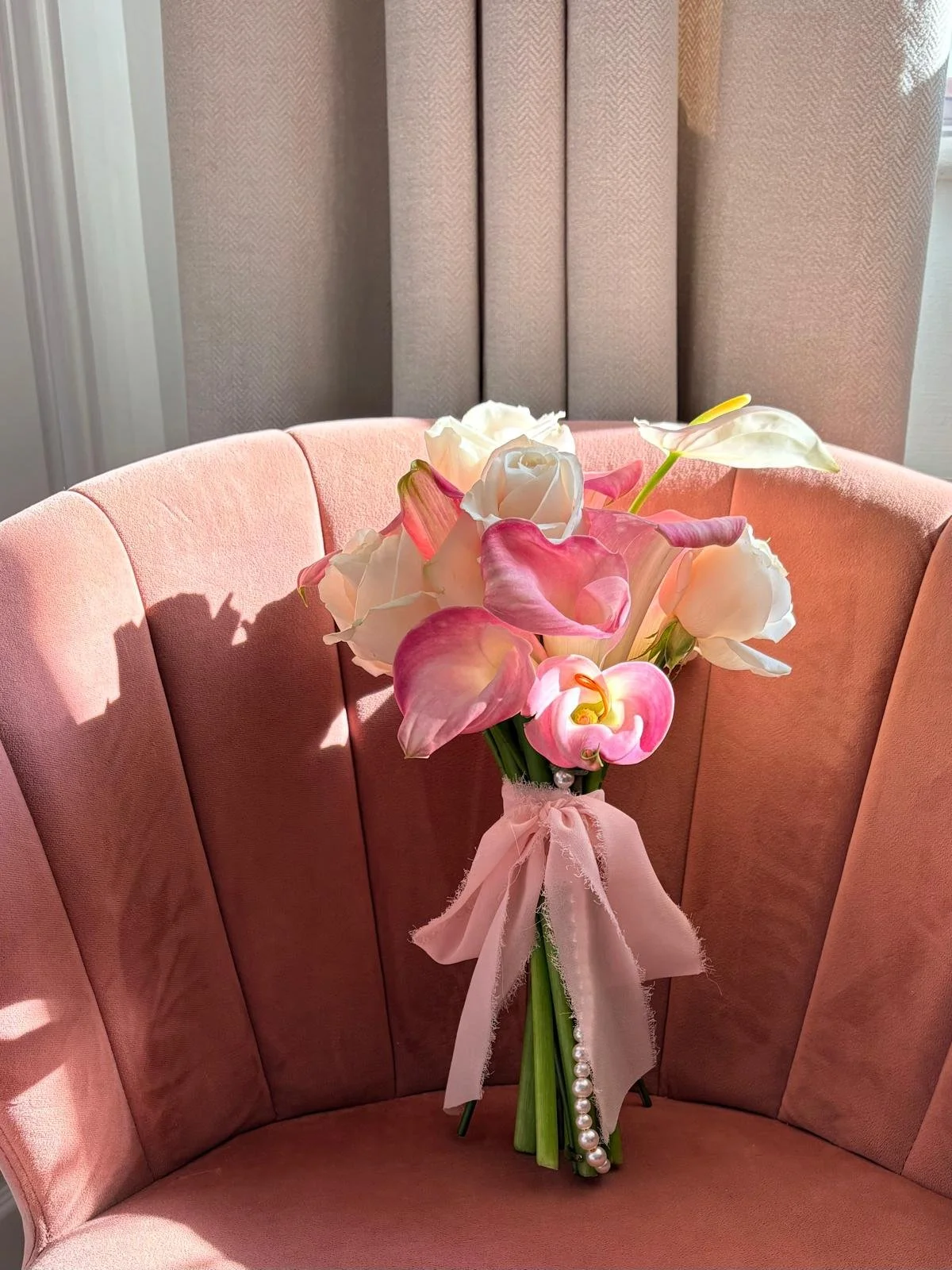 A bouquet of pink and white flowers, including calla lilies and roses, tied with a pink ribbon, resting on a pink upholstered chair with curtains in the background.