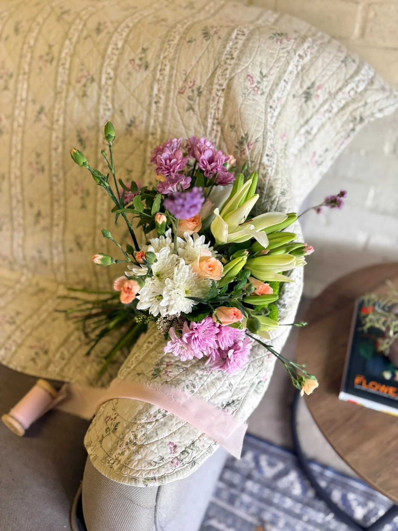 A bouquet of mixed flowers, including white lilies, pink carnations, and purple and white chrysanthemums, resting on a floral-patterned quilted fabric.
