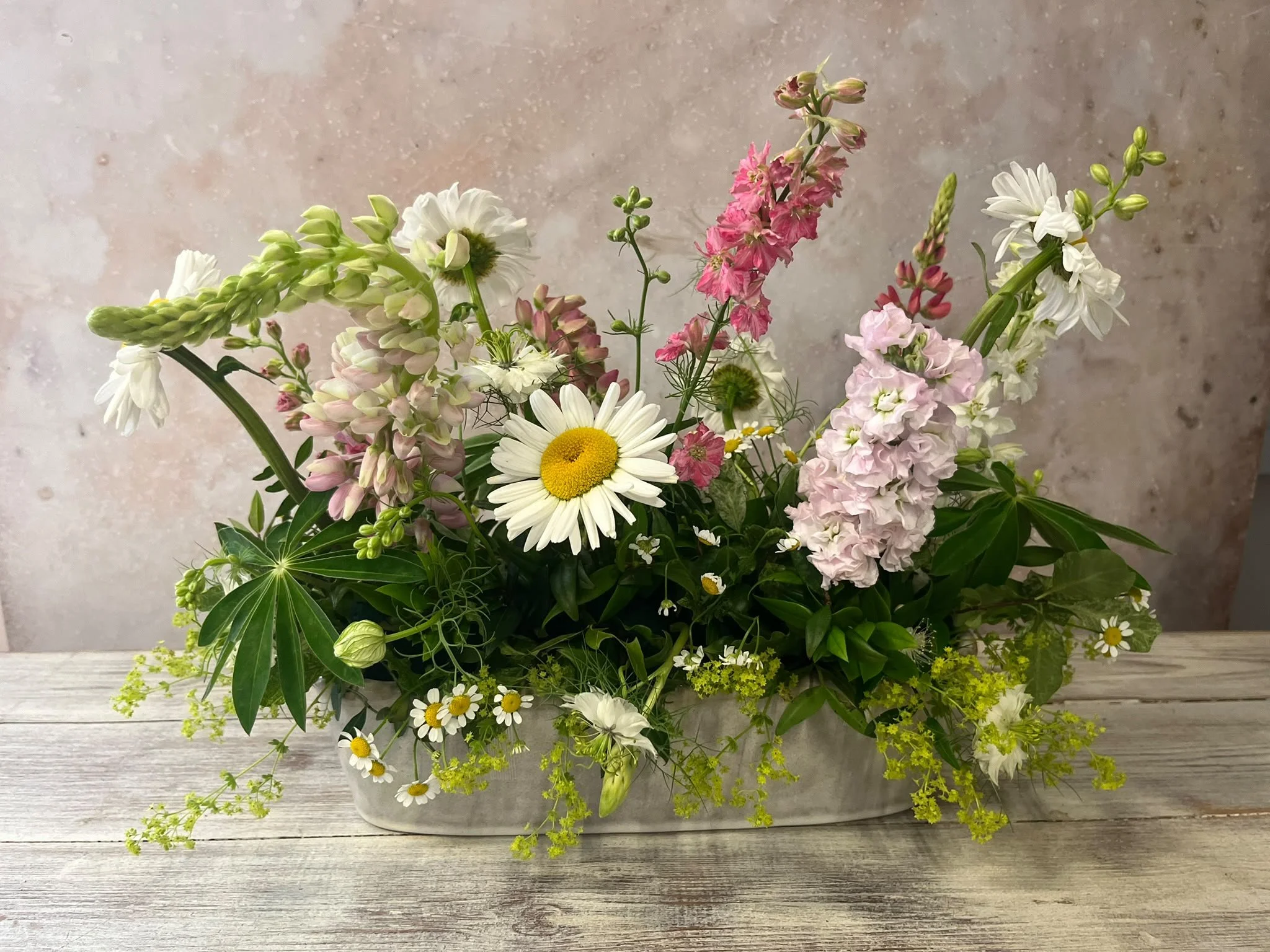 A floral arrangement in a white container on a wooden surface, featuring daisies, lilies, snapdragons, and assorted greenery.