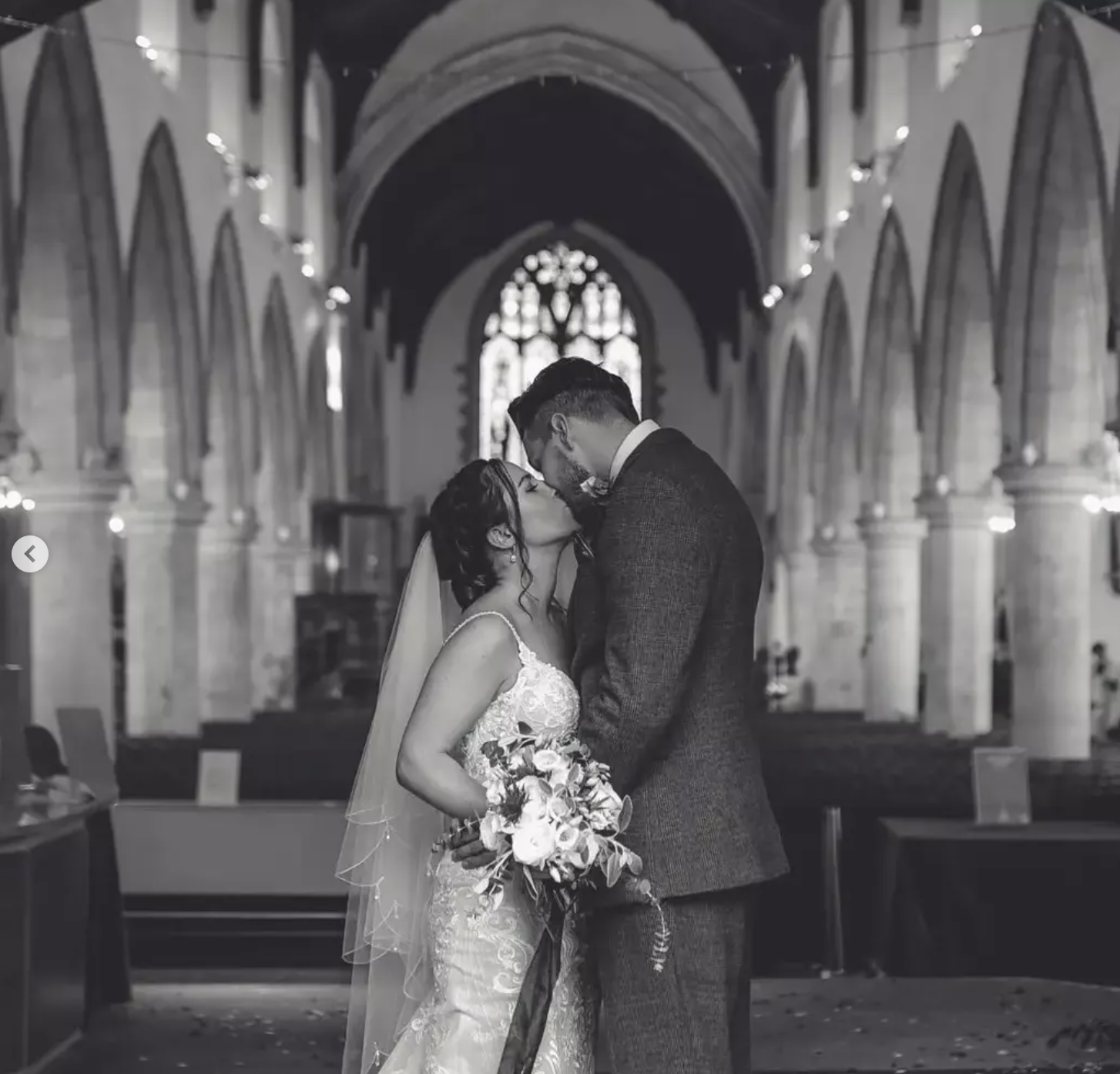 A black-and-white photograph of a bride and groom sharing a kiss in a church with tall stained glass windows and arched architecture, the bride holding a bouquet of flowers and wearing a lace wedding dress with a veil, the groom in a suit.