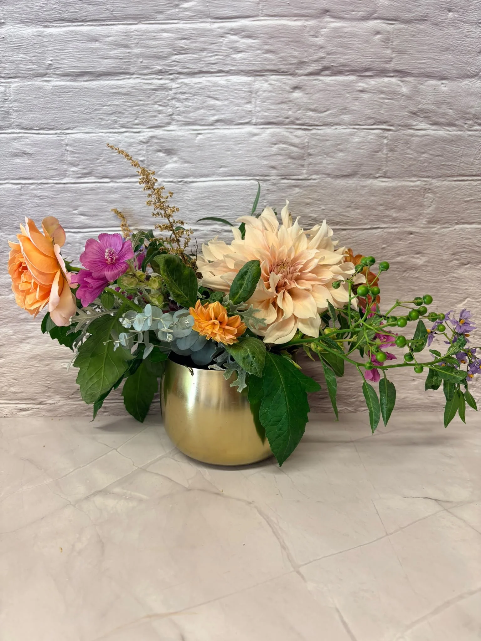 A floral arrangement in a gold vase with orange, pink, peach, and purple flowers and green foliage against a white brick wall background.
