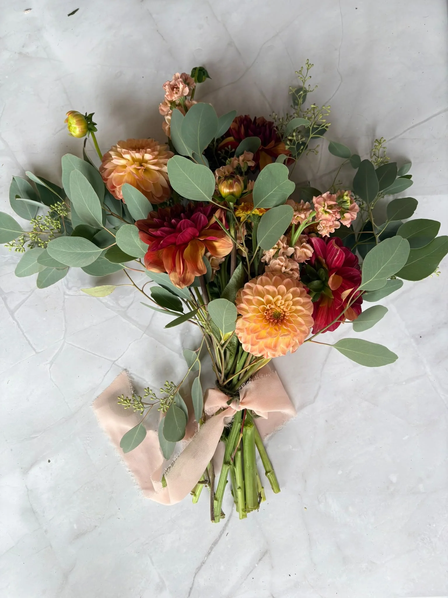 A bouquet of pink, orange, and red flowers with green eucalyptus leaves wrapped with a light pink ribbon, resting on a light-colored marble surface.