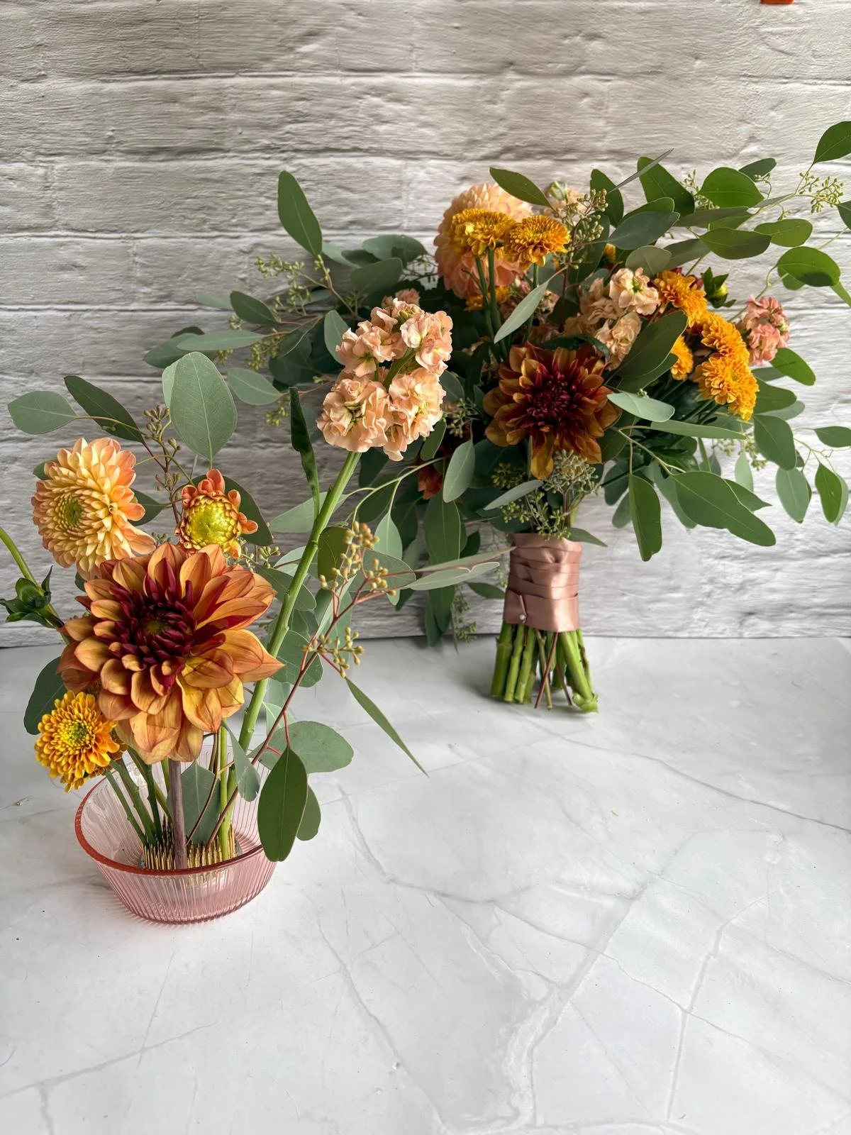 Two bouquets of mixed flowers and greenery on a white surface against a textured white wooden wall. One bouquet is in a pink vase, the other is wrapped with brown ribbon.
