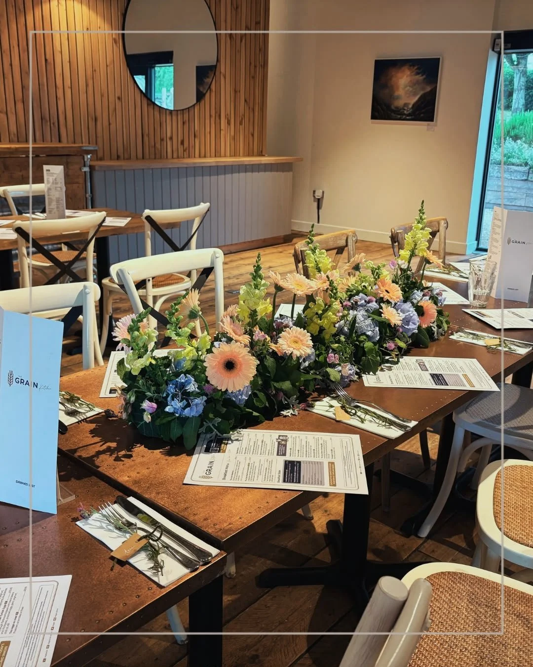 Table decorated with a large floral centerpiece featuring pink, yellow, and purple flowers in a restaurant or cafe.