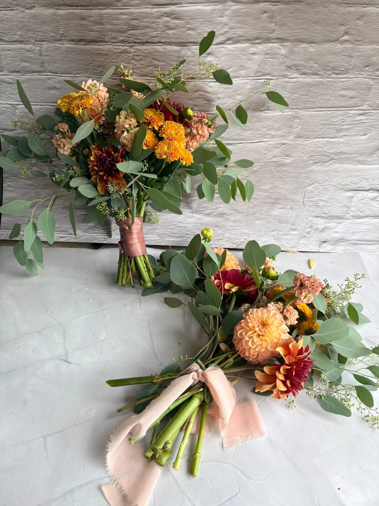 Two bouquets of colorful flowers with green leaves and pink ribbons on a white textured surface, with a white wooden wall in the background.