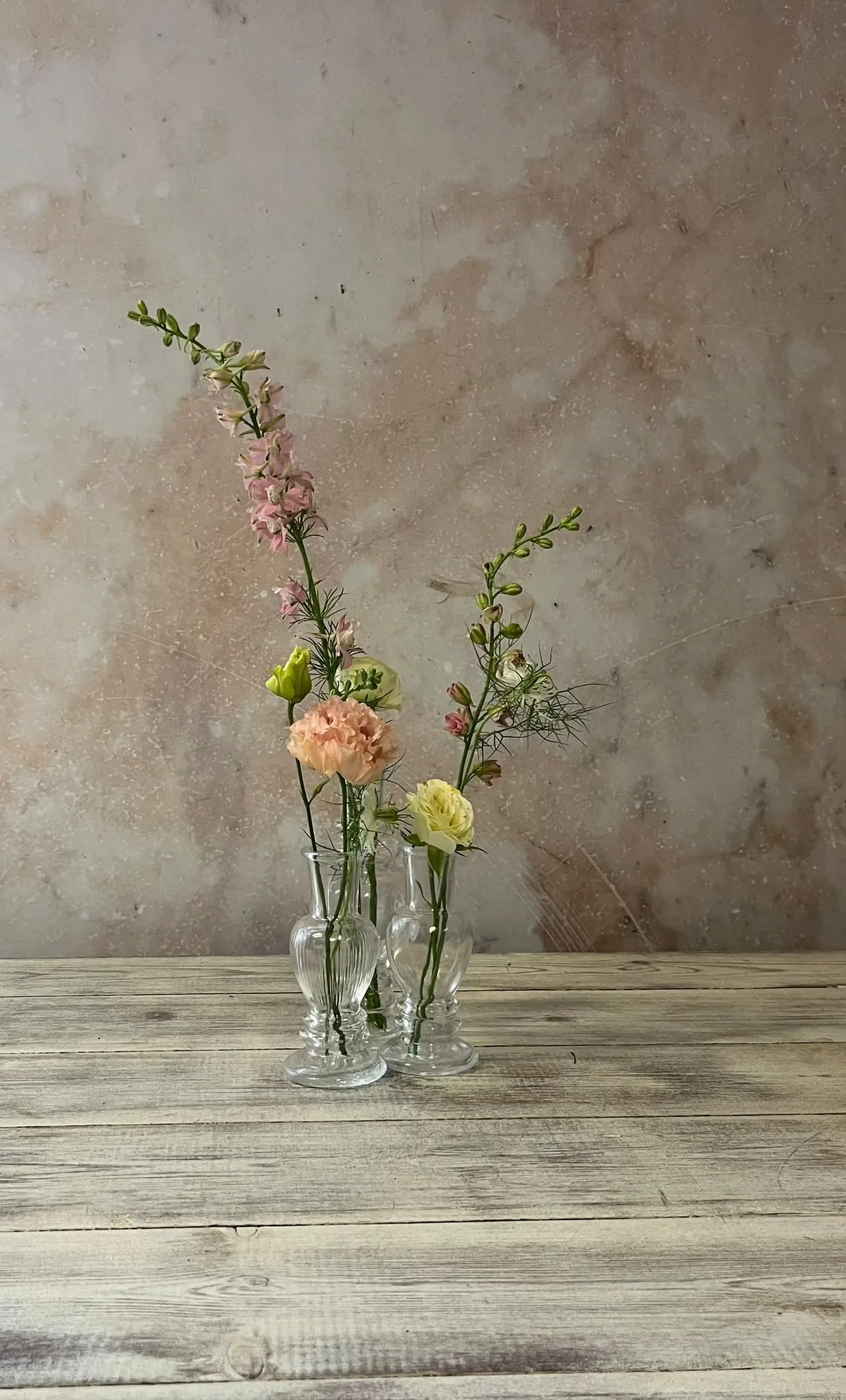 Three glass vases with various pink, yellow, and white flowers on a wooden surface against a textured beige wall.