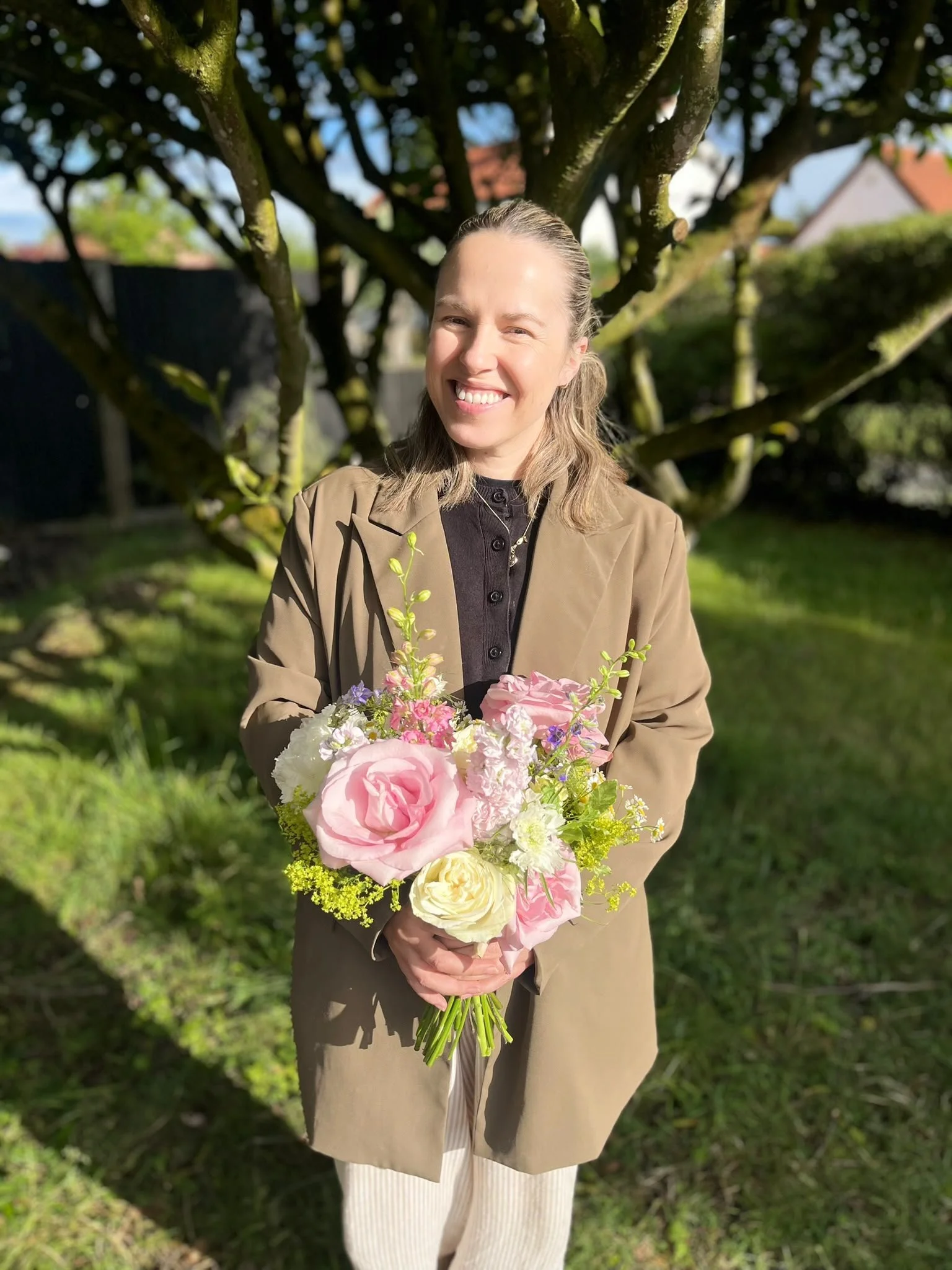 A woman with blonde hair smiling while holding a bouquet of pink, white, and purple flowers outdoors during daylight.