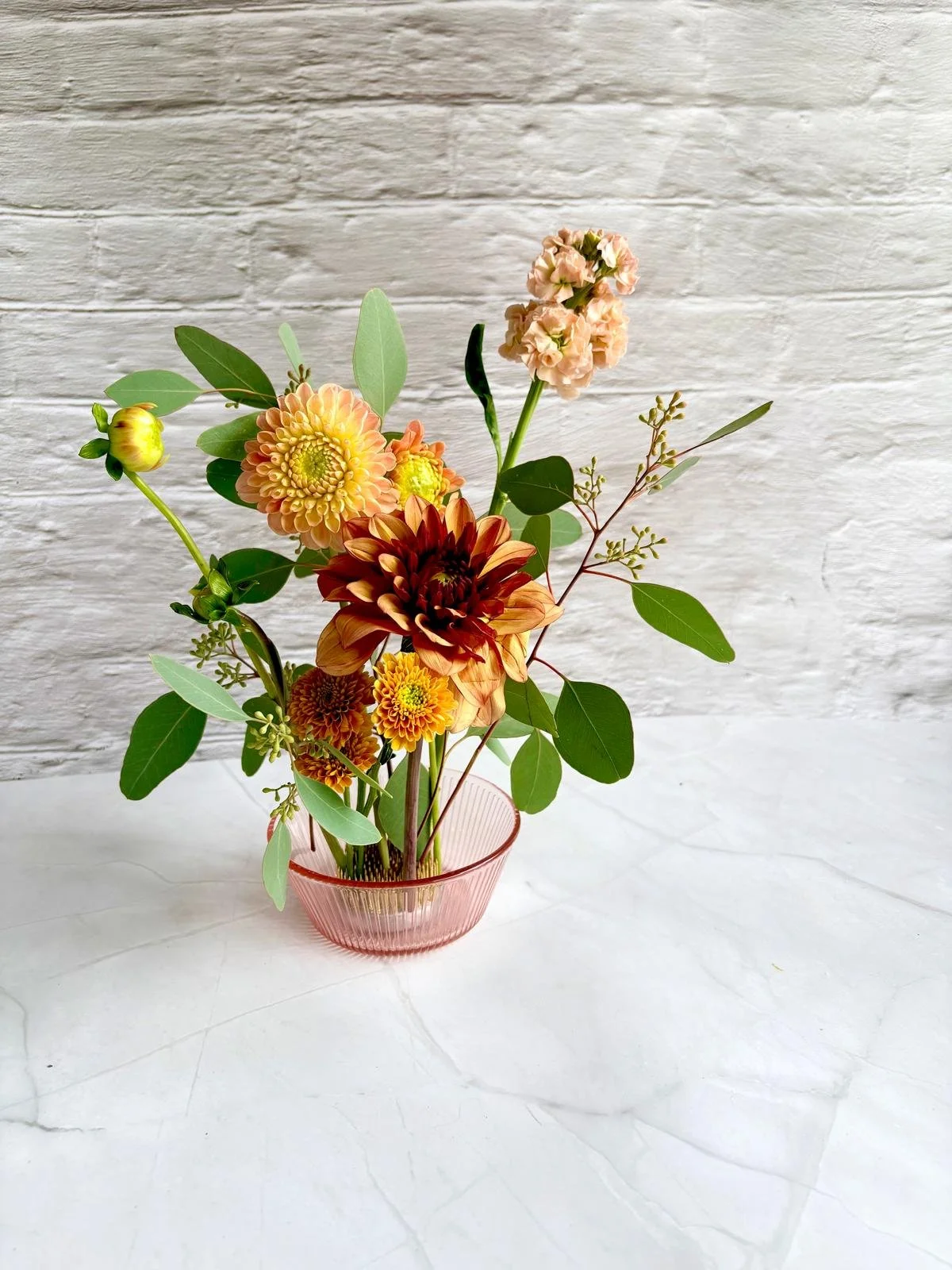 Arrangement of various flowers and green leaves in a pink glass vase, set against a white brick wall background.