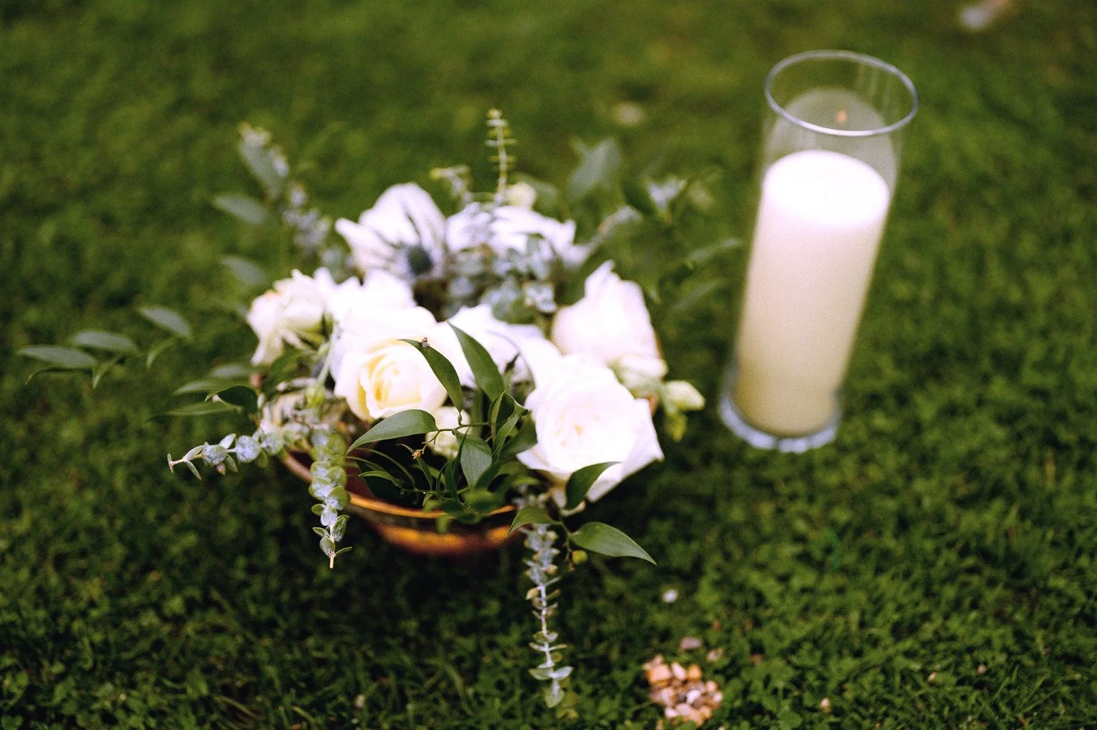 A floral arrangement with white roses and greenery in a small basket, next to a tall glass candle holder with a white candle inside, on a green grass background.