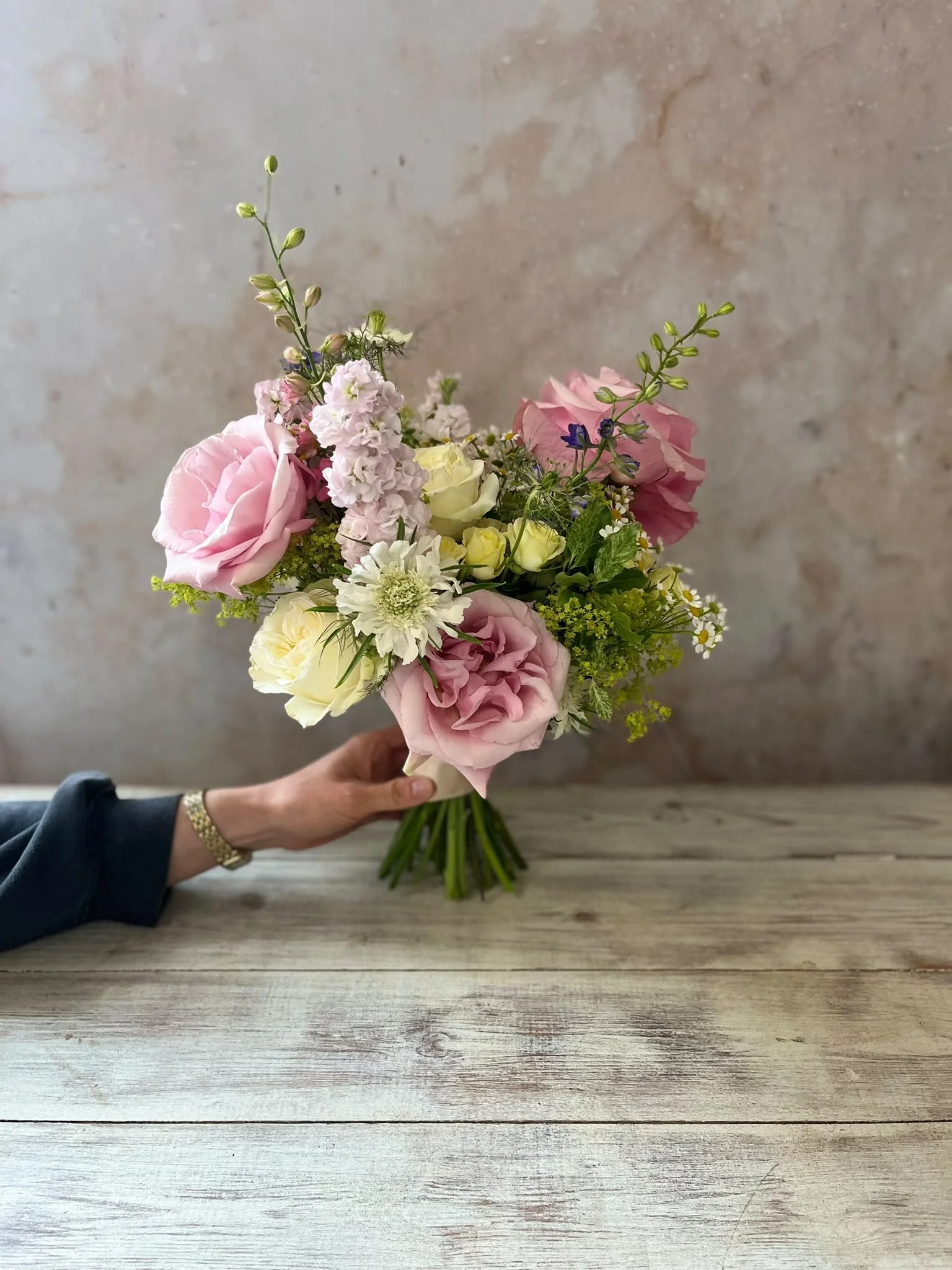 A person holding a bouquet of pink, white, and purple flowers against a rustic wooden surface with a plain background.