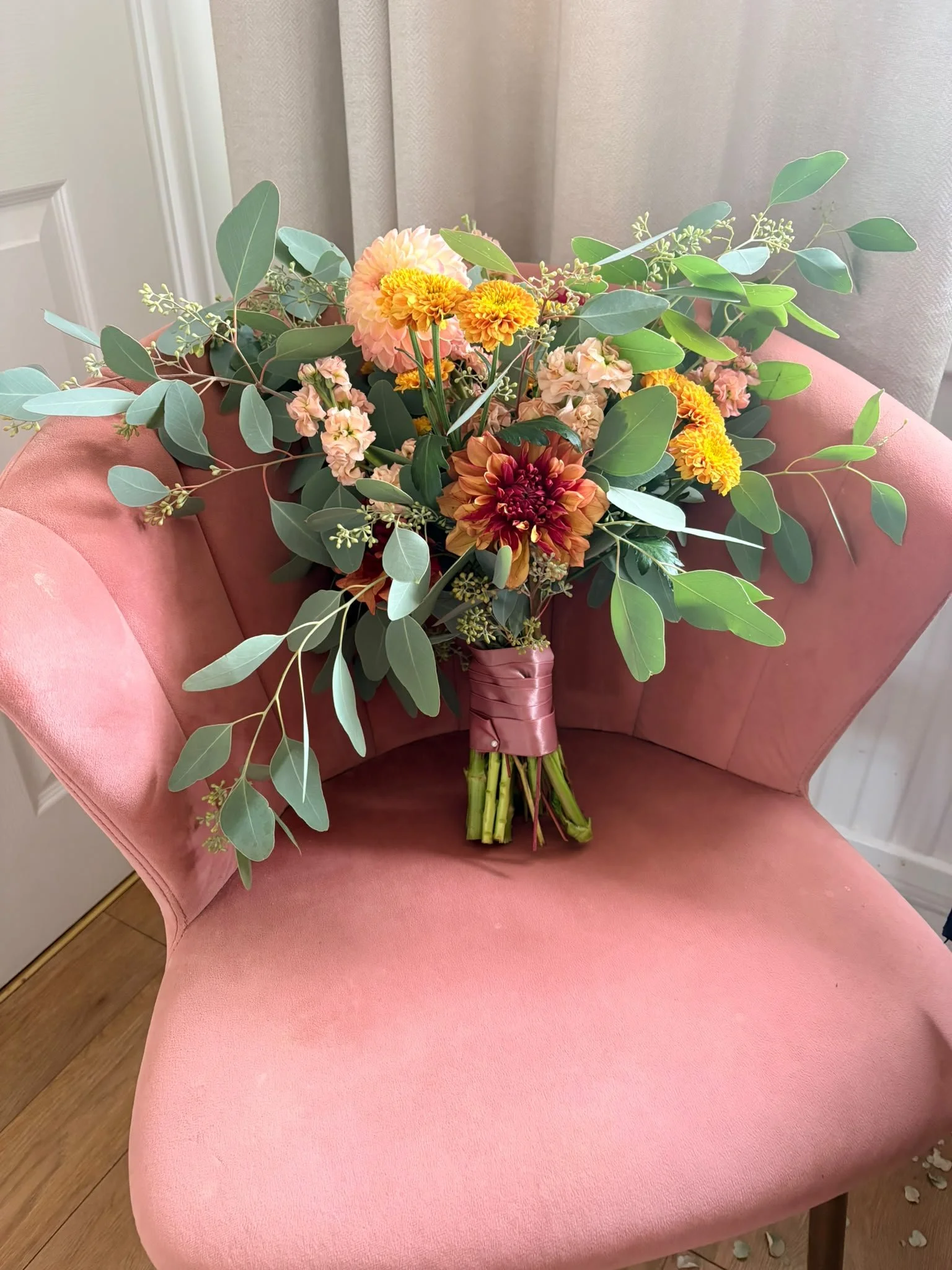 A bouquet of colorful flowers including pink, yellow, and red blooms with green eucalyptus and other foliage resting on a pink upholstered chair near a window with beige curtains.