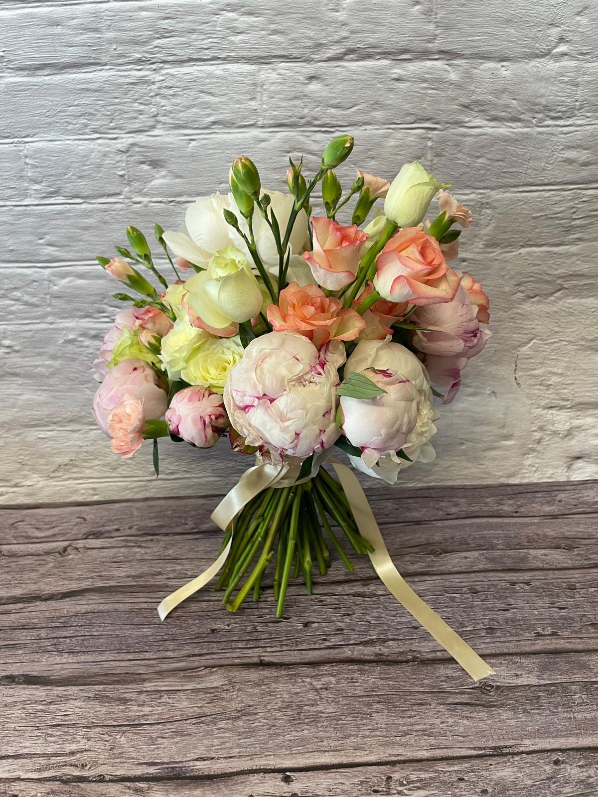 A bouquet of white, pink, and peach roses and peonies tied with a yellow ribbon, placed on a wooden surface against a white brick wall.