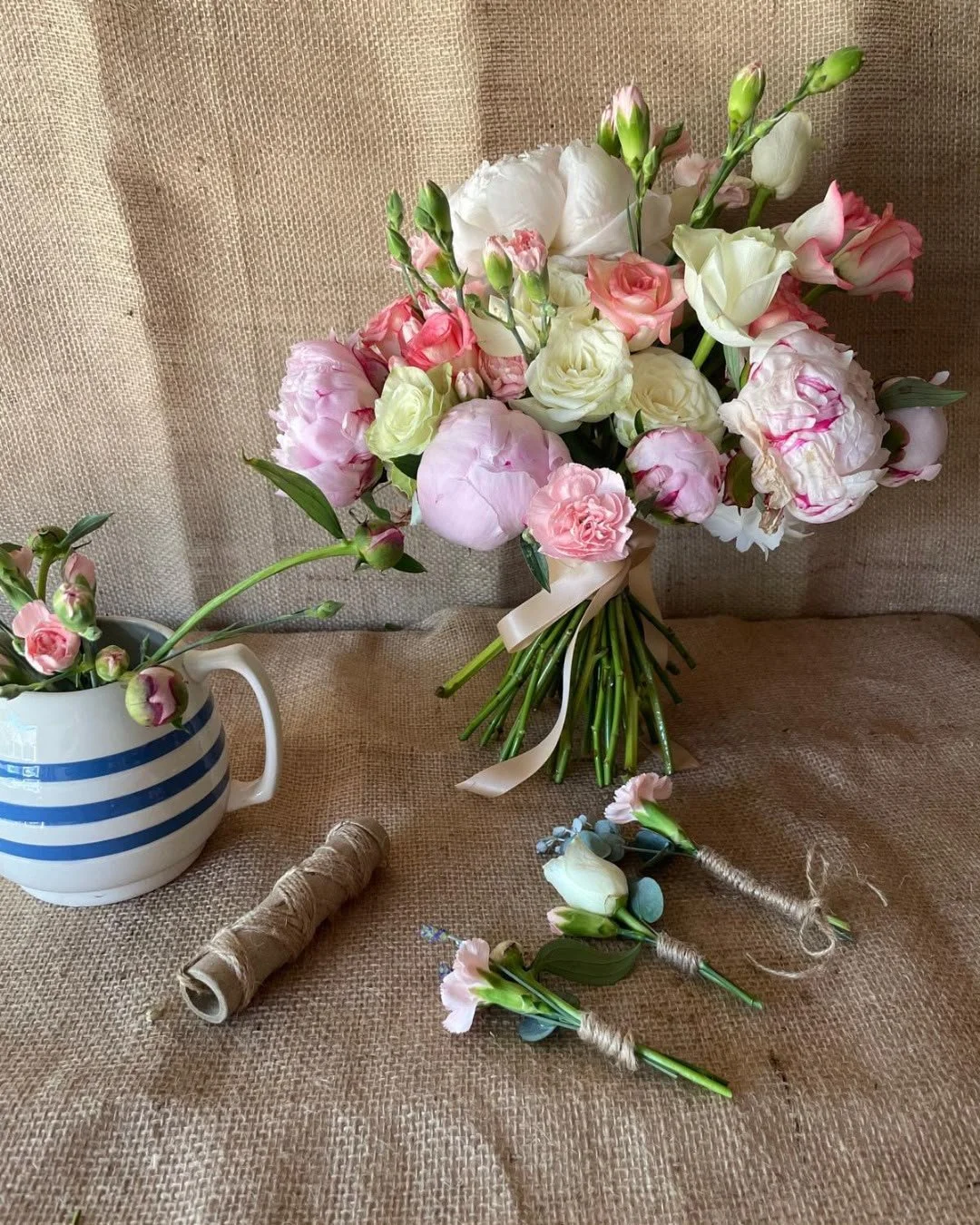 A bouquet of pink and white flowers with some buds, a spool of twine, and a small floral arrangement on a brown burlap surface.