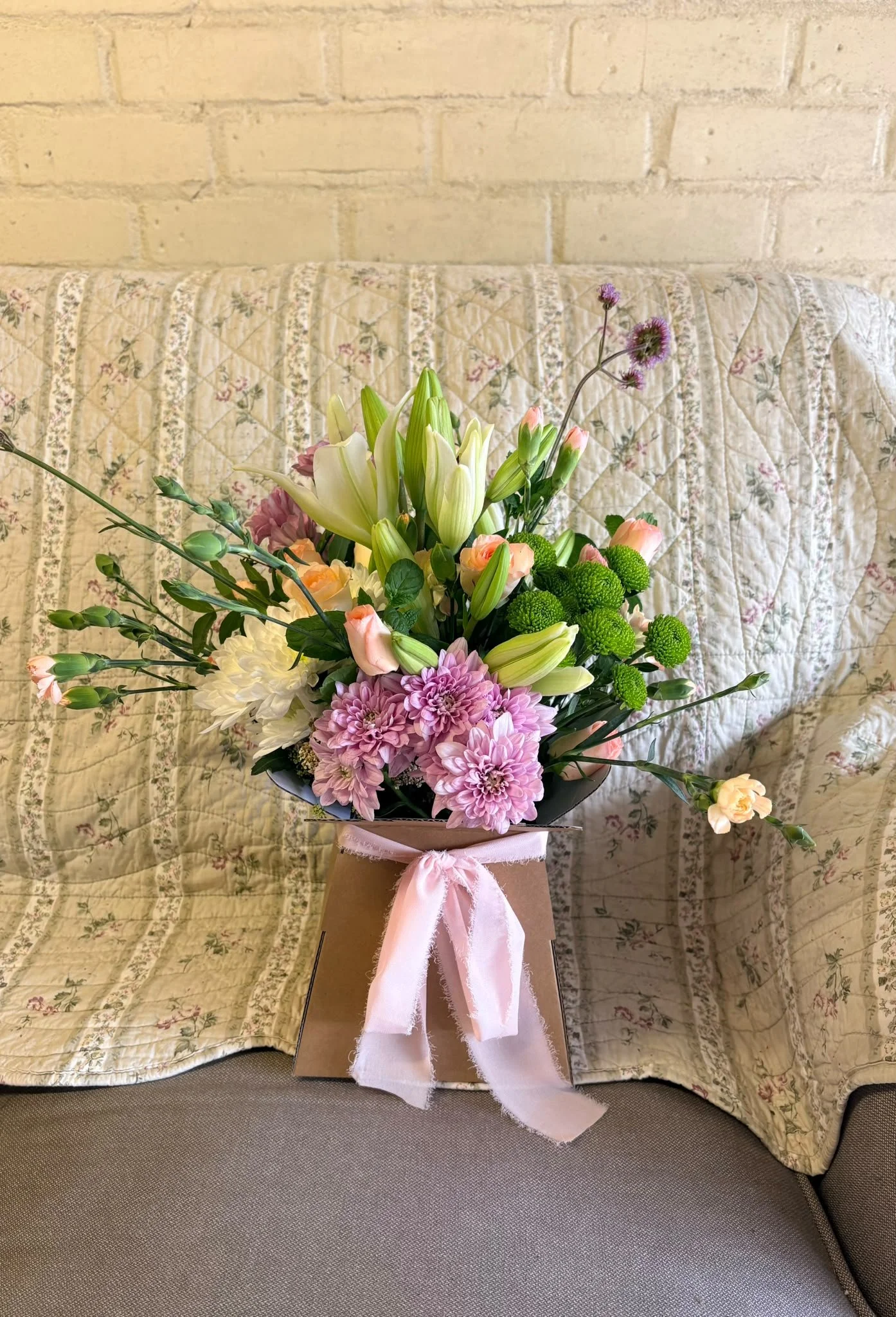 A bouquet of mixed flowers in a brown paper bag tied with pink ribbon, placed on a gray couch with floral quilted fabric in the background.