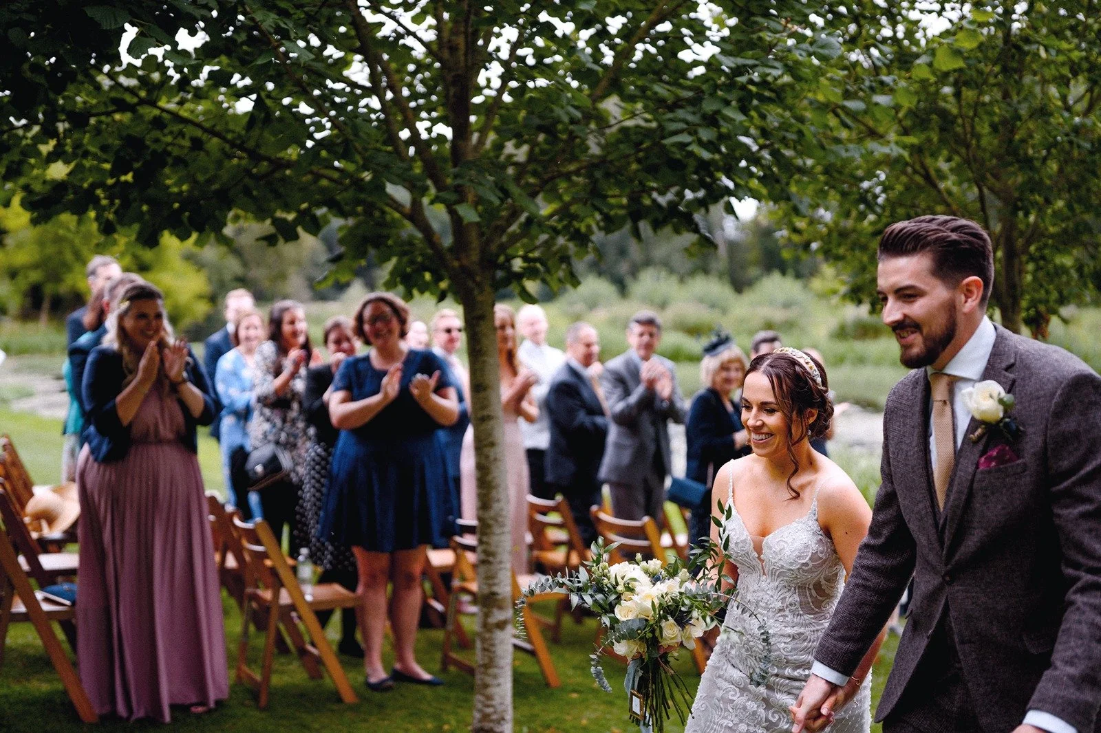 A bride and groom walking hand in hand down an outdoor aisle during a wedding ceremony, with friends and family clapping and smiling in the background under trees.