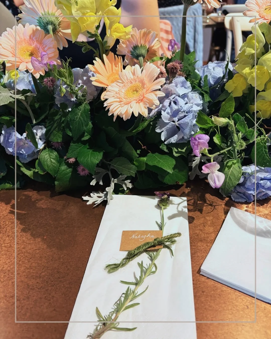 A floral arrangement with pink gerbera daisies, blue hydrangeas, and yellow snapdragons, with greenery, and a sprig of rosemary on a white envelope labeled 'Nataoha' on a brown table.