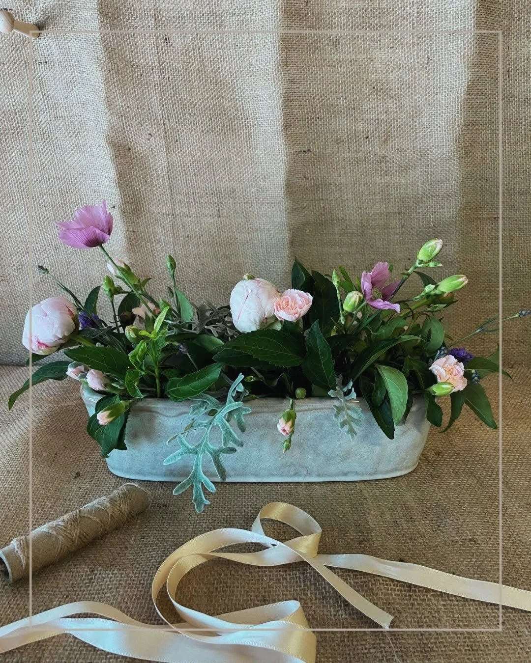 A floral arrangement in a white oval ceramic vase with pink and purple flowers, green leaves, and decorative gray foliage, set on a brown textured surface with a spool of twine and white ribbon in the foreground, against a beige fabric background.