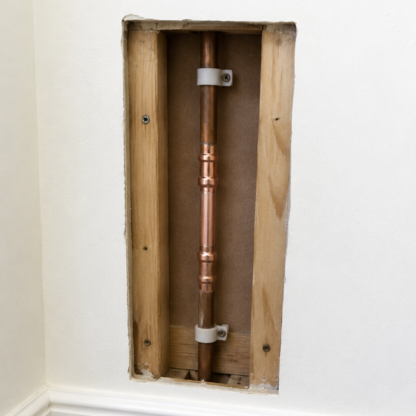 A copper plumbing pipe installed in a wall opening with wooden framing and white wall casing.