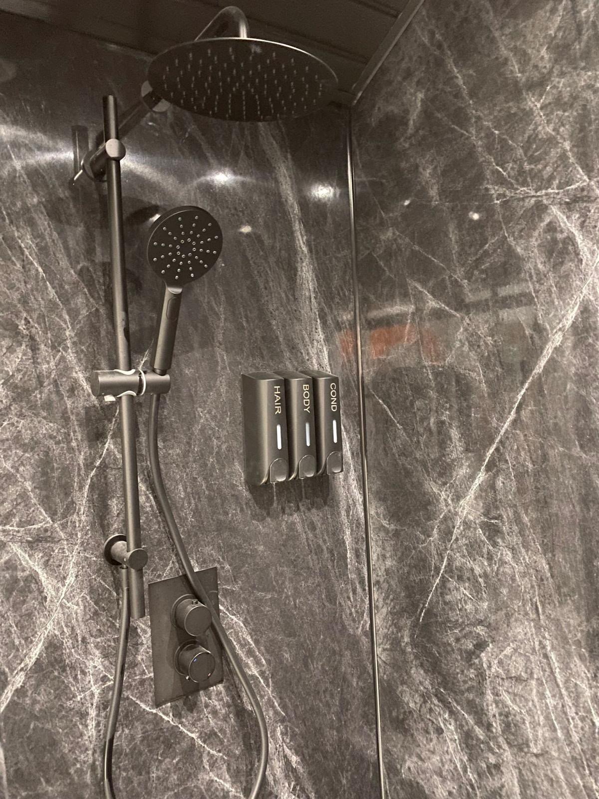 Shower with gray marble walls, a rain showerhead, a handheld showerhead, and three labeled dispensers for hair, body, and sound.