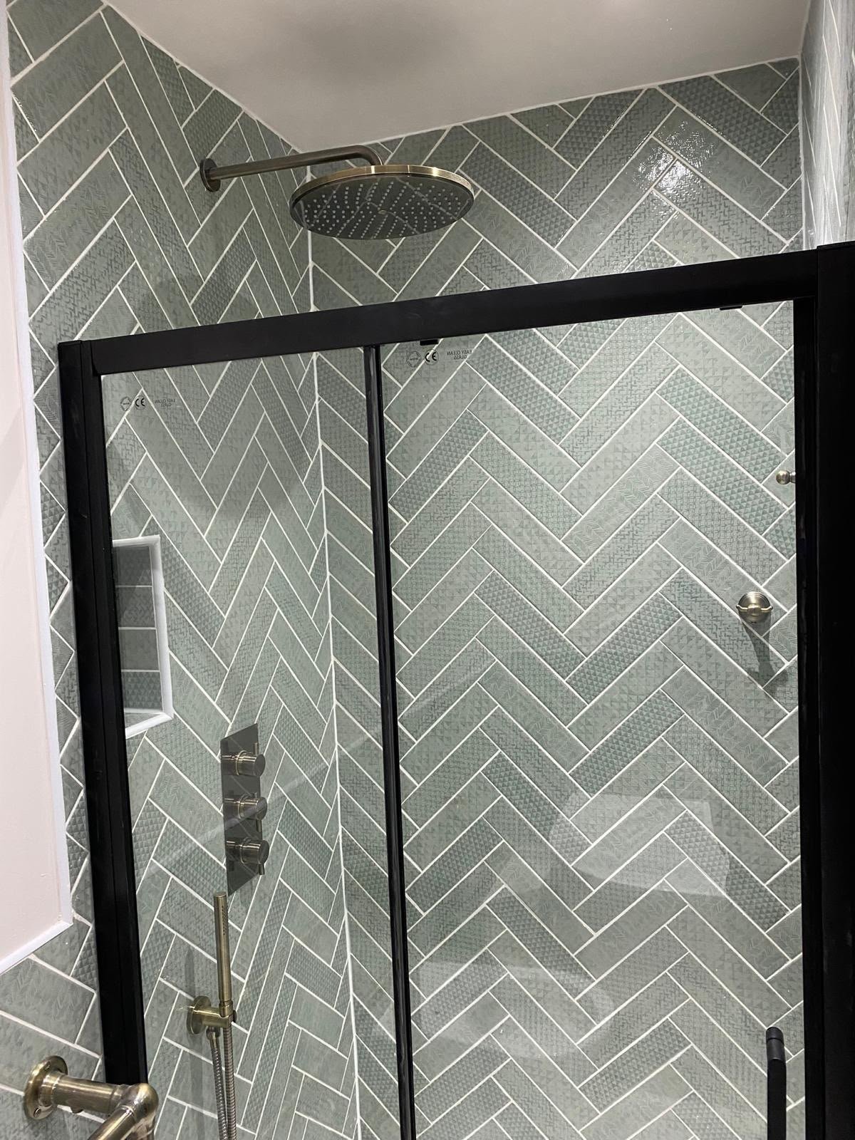 Shower with gray herringbone-patterned tile walls, black-framed glass door, large round showerhead, and built-in wall niche.