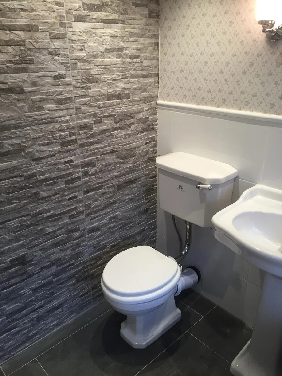 A small bathroom with gray stone-textured wall tiles on one side and floral wallpaper on the other. It contains a white toilet and a white sink with a curved edge, with a wall-mounted light above.