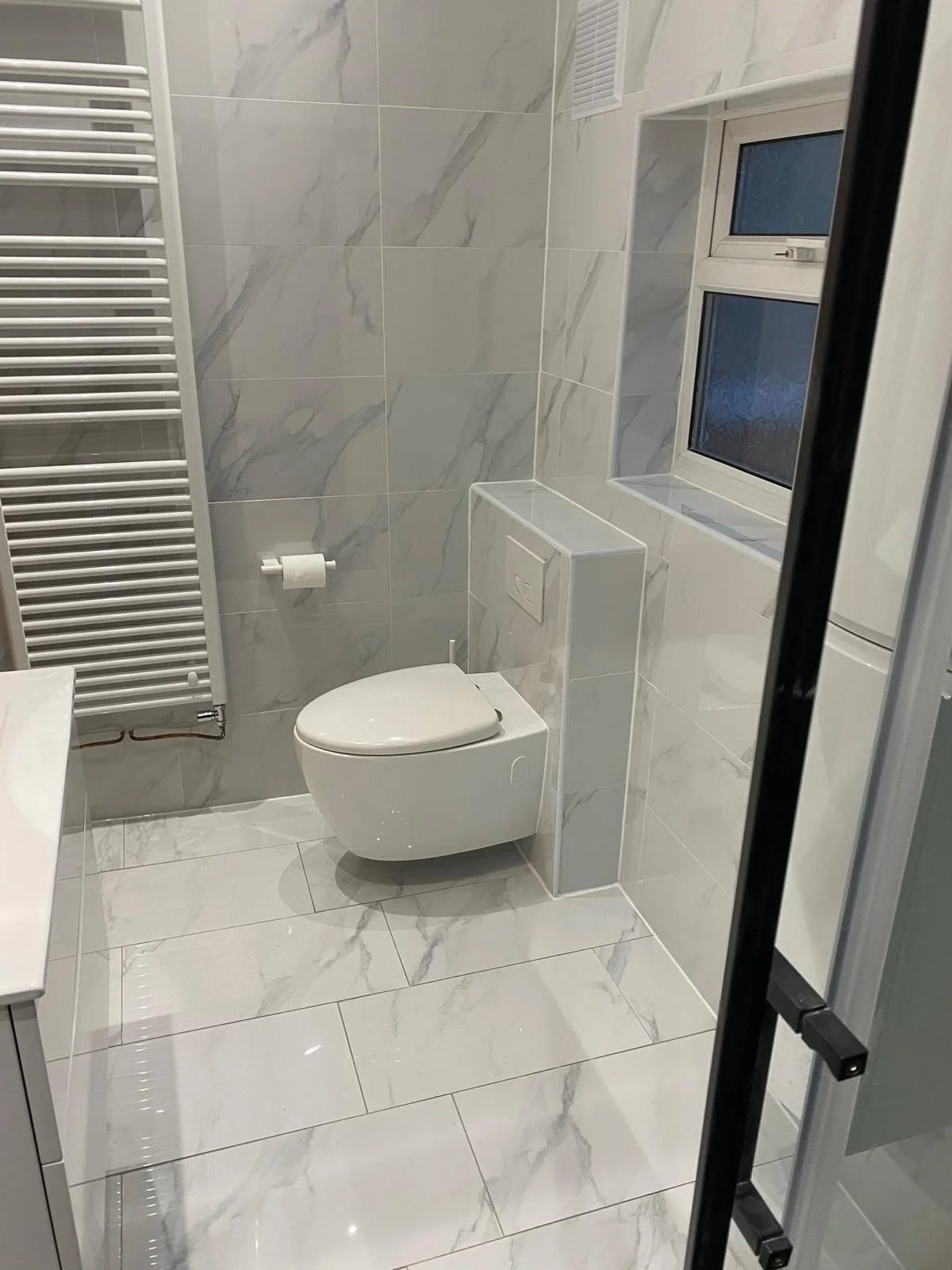 A small modern bathroom with white marble tiles on the walls and floor. It has a wall-mounted toilet, a window, a radiator towel warmer, and a flush panel. The photo is taken through a glass shower door.