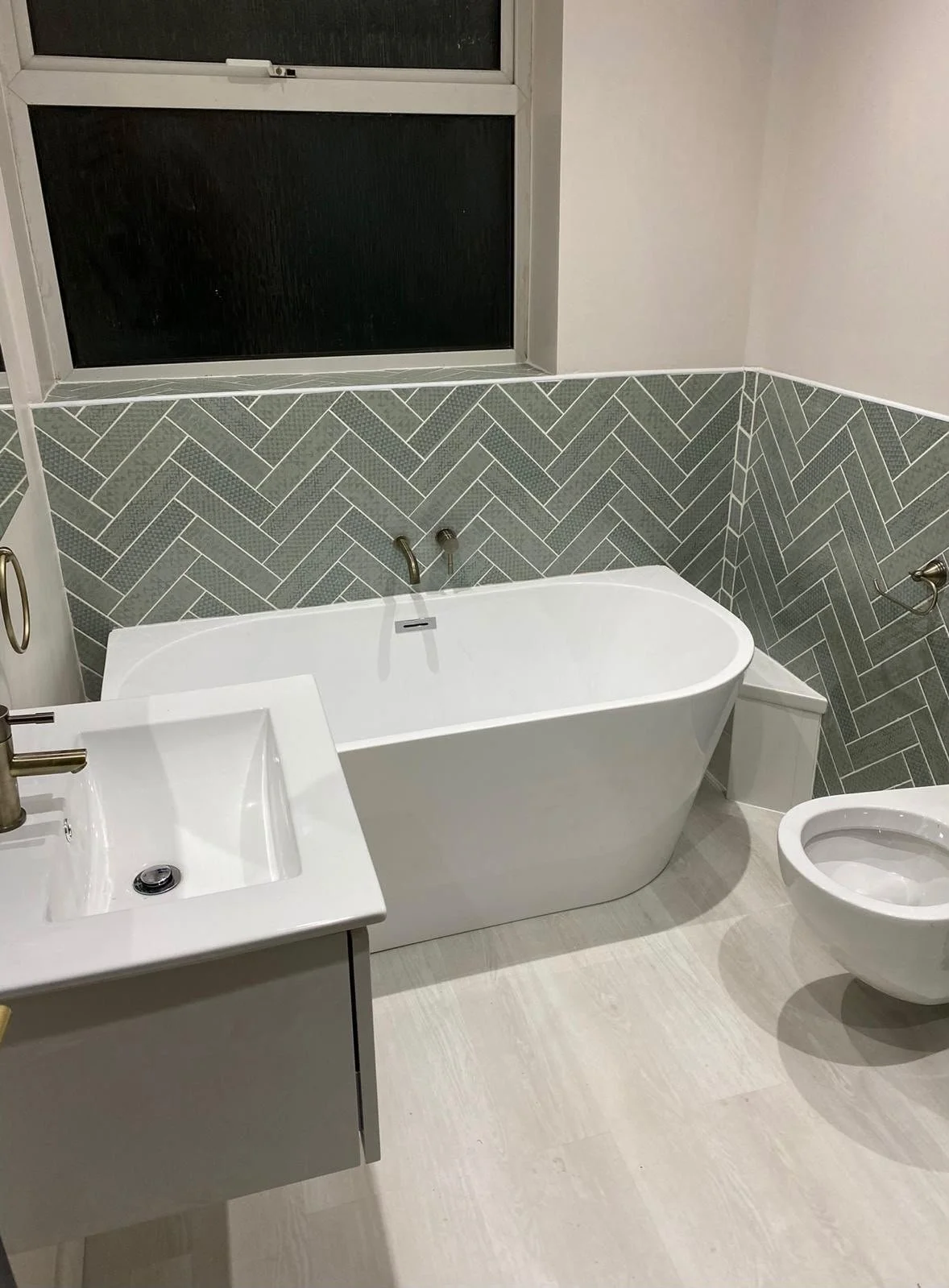 Modern bathroom with a freestanding bathtub, a small white vanity with a sink, a toilet, and a window with frosted glass. The walls feature a gray herringbone tile pattern halfway up.