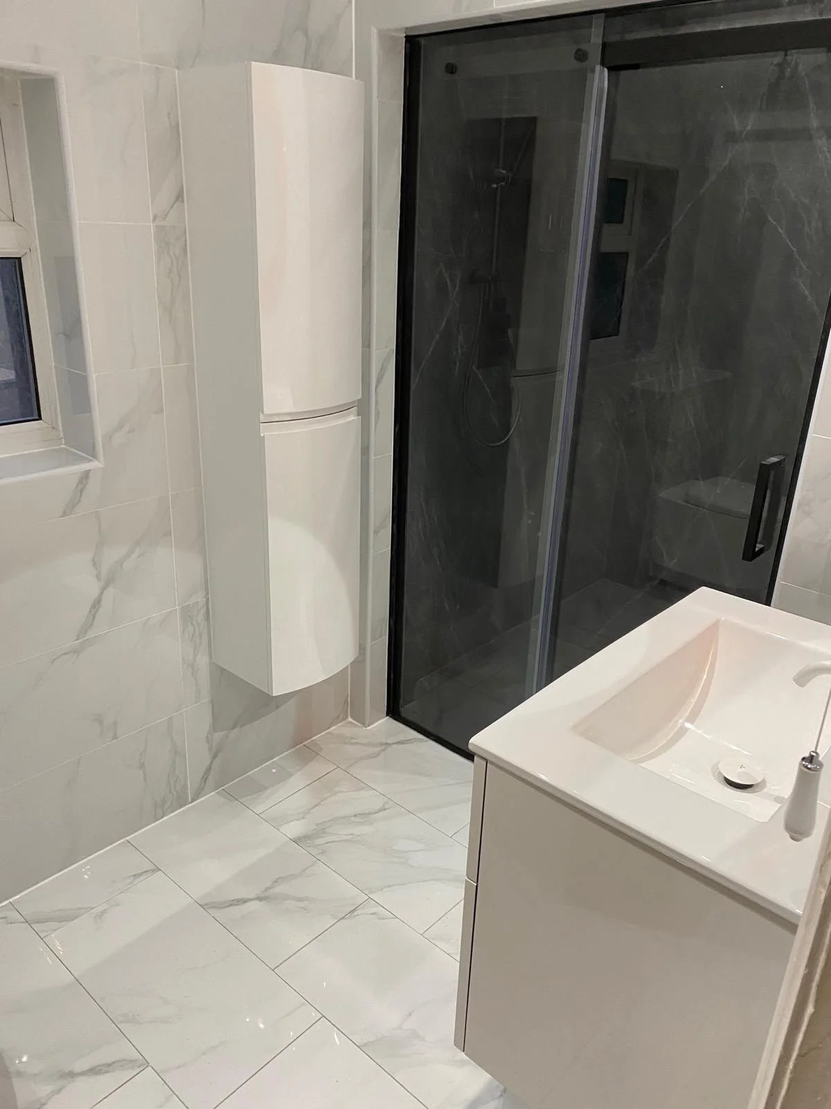 Modern bathroom with white marble tiles, a white vanity with an integrated sink, a black shower enclosure with a glass door, and a small window.