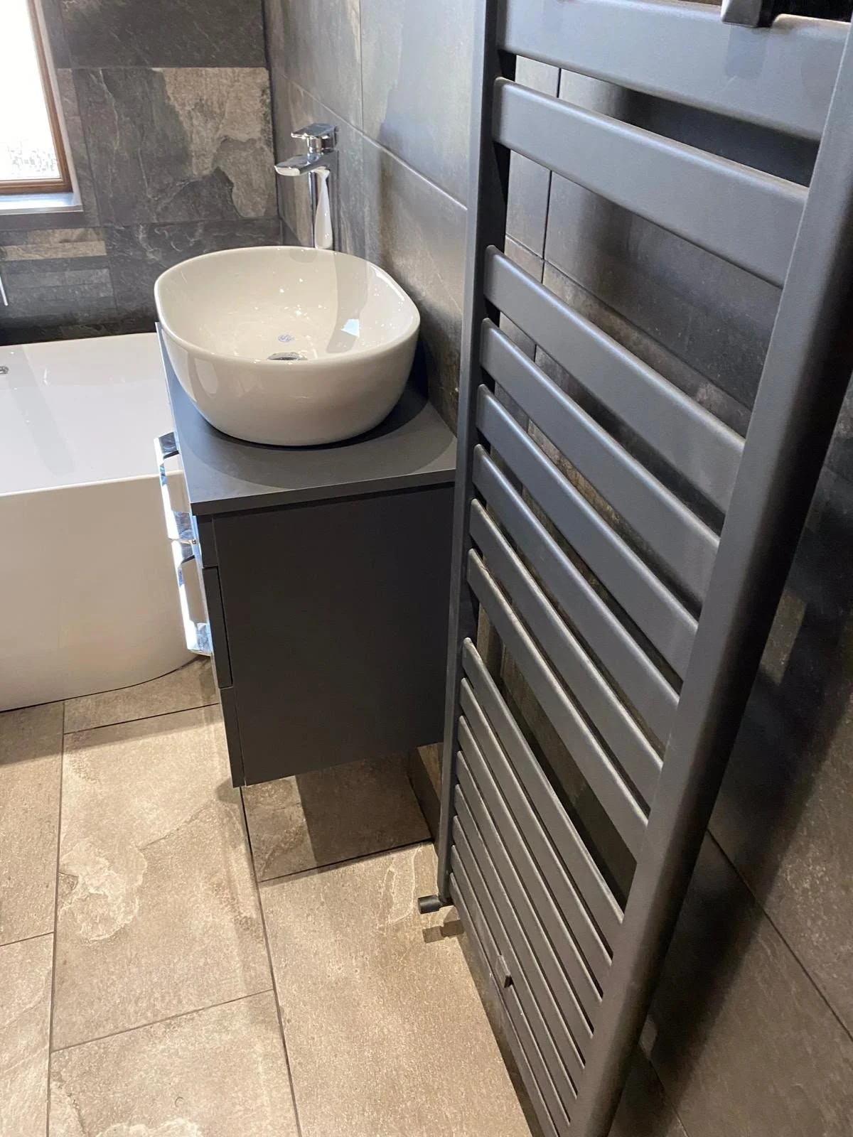Modern bathroom with a white vessel sink on a gray vanity, a black towel radiator, and gray tiled walls and floor.