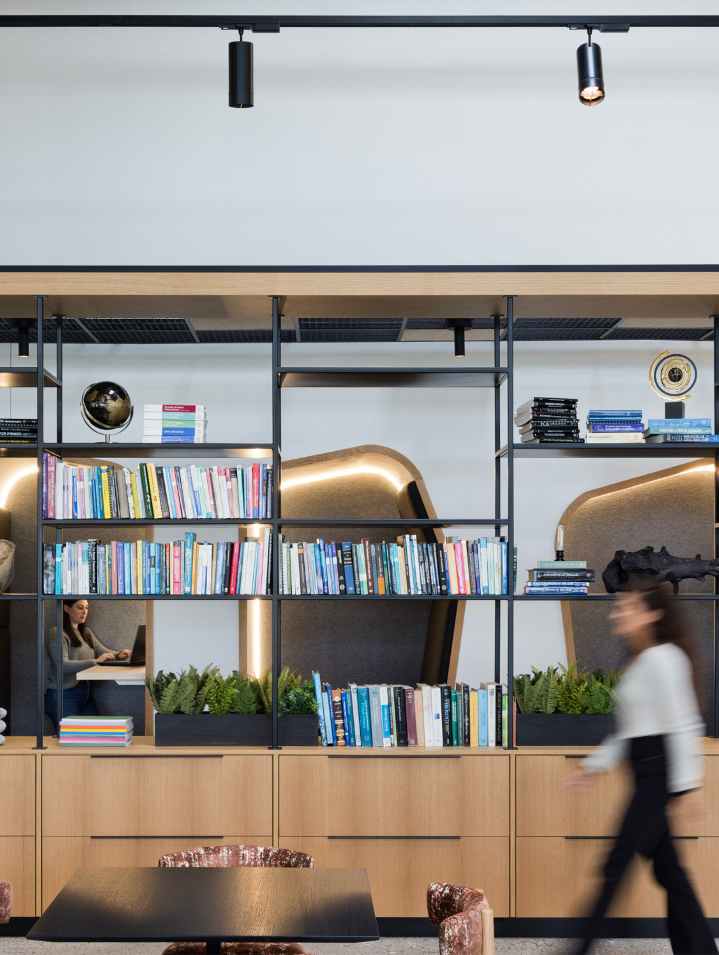 Interior view of a modern office space with a black bookshelf filled with colorful books, a small globe, and decorative plants, with two women working at desks in the background and a person walking past in the foreground.