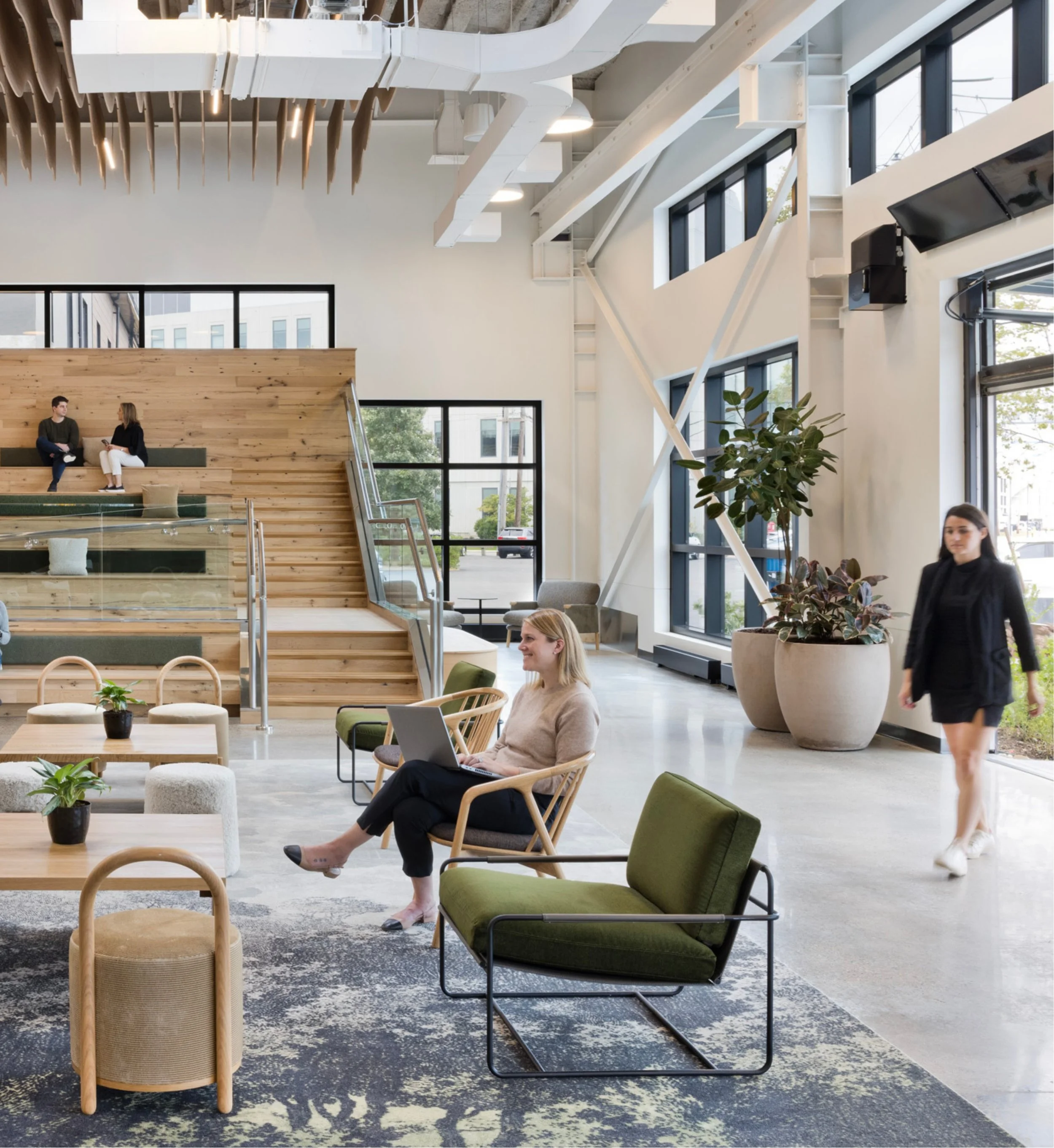 Modern lobby interior with large windows, green plants, and various seating arrangements. Several people are engaging in different activities, including a woman sitting with a laptop, two people talking on a staircase, and a woman walking through the space. Wooden accents and contemporary design elements are visible.