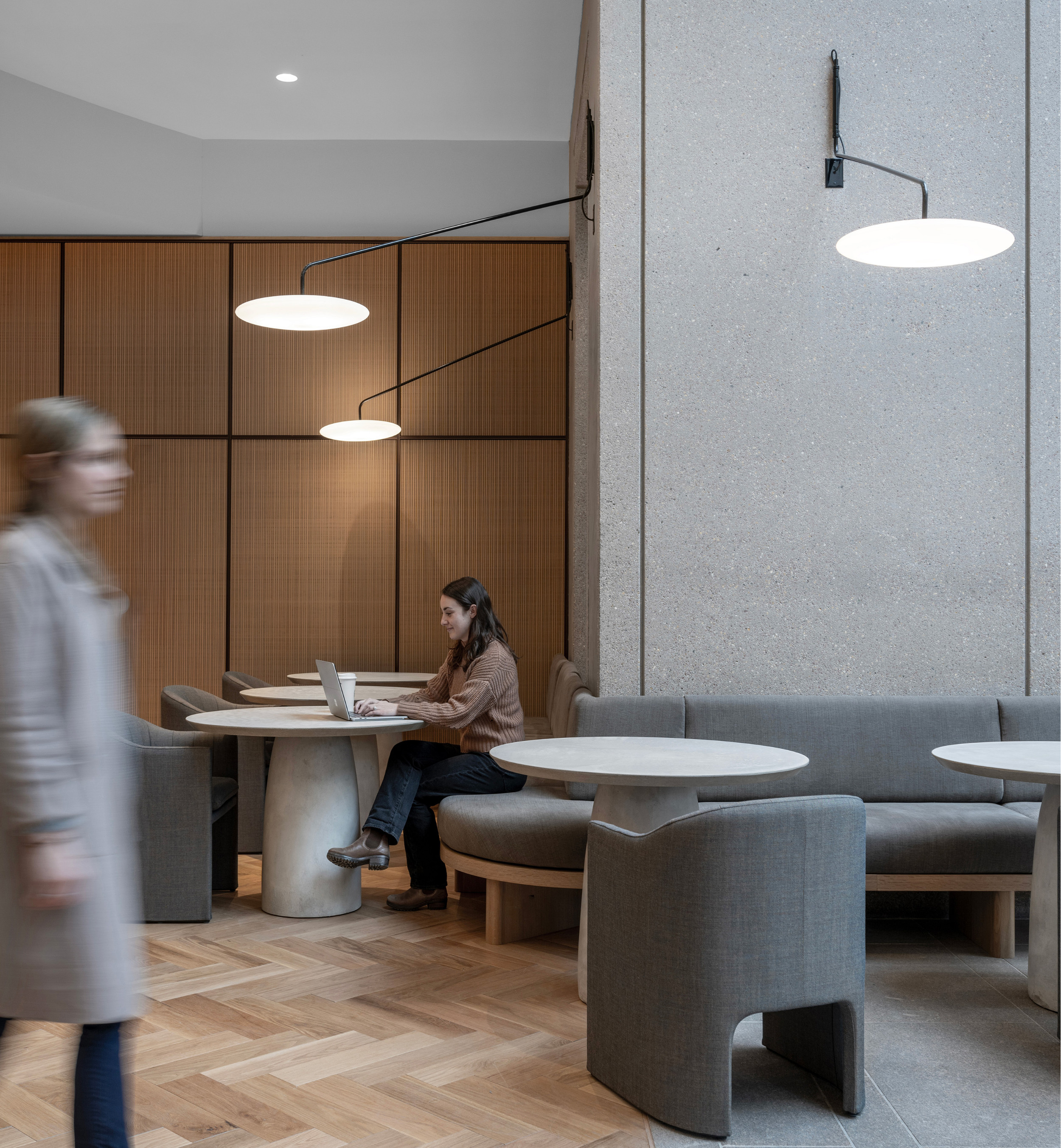 Urban cafe interior with round tables, cushioned seating, wooden wall, concrete wall, modern lighting, and two women using laptops.