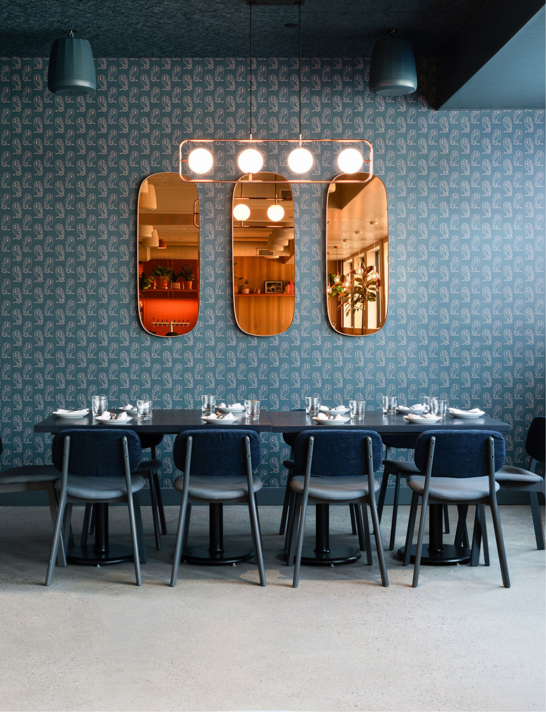 A modern dining room with a long black table set with white plates, glasses, and utensils, surrounded by blue chairs. The background features a textured wall with a repeating pattern of abstract shapes, a set of three oval mirrors, and a decorative light fixture with four bright bulbs.