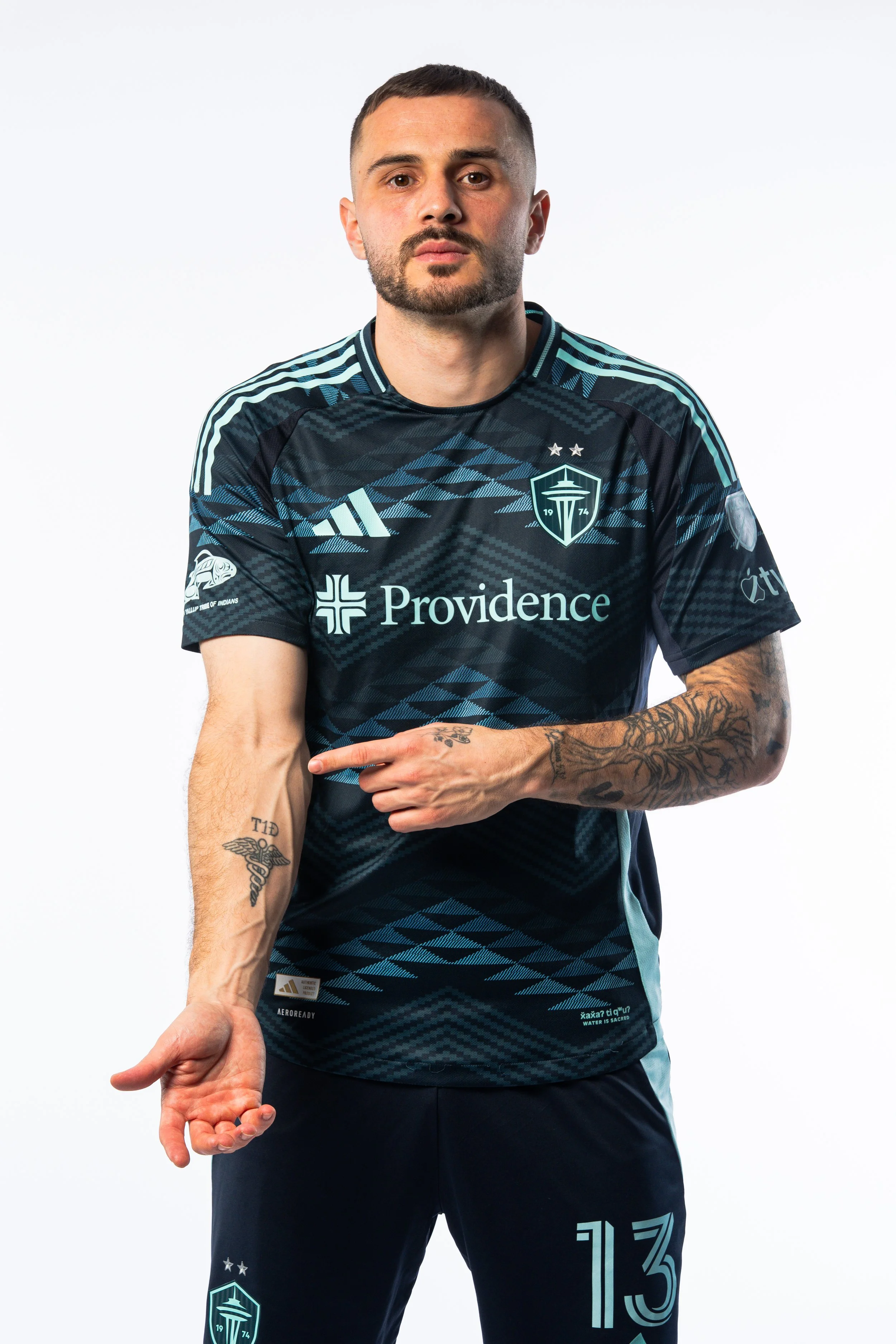 A man with tattoos on his arms and neck wearing a black and blue Seattle Sounders soccer jersey and black pants, gesturing with his right hand against a plain white background.