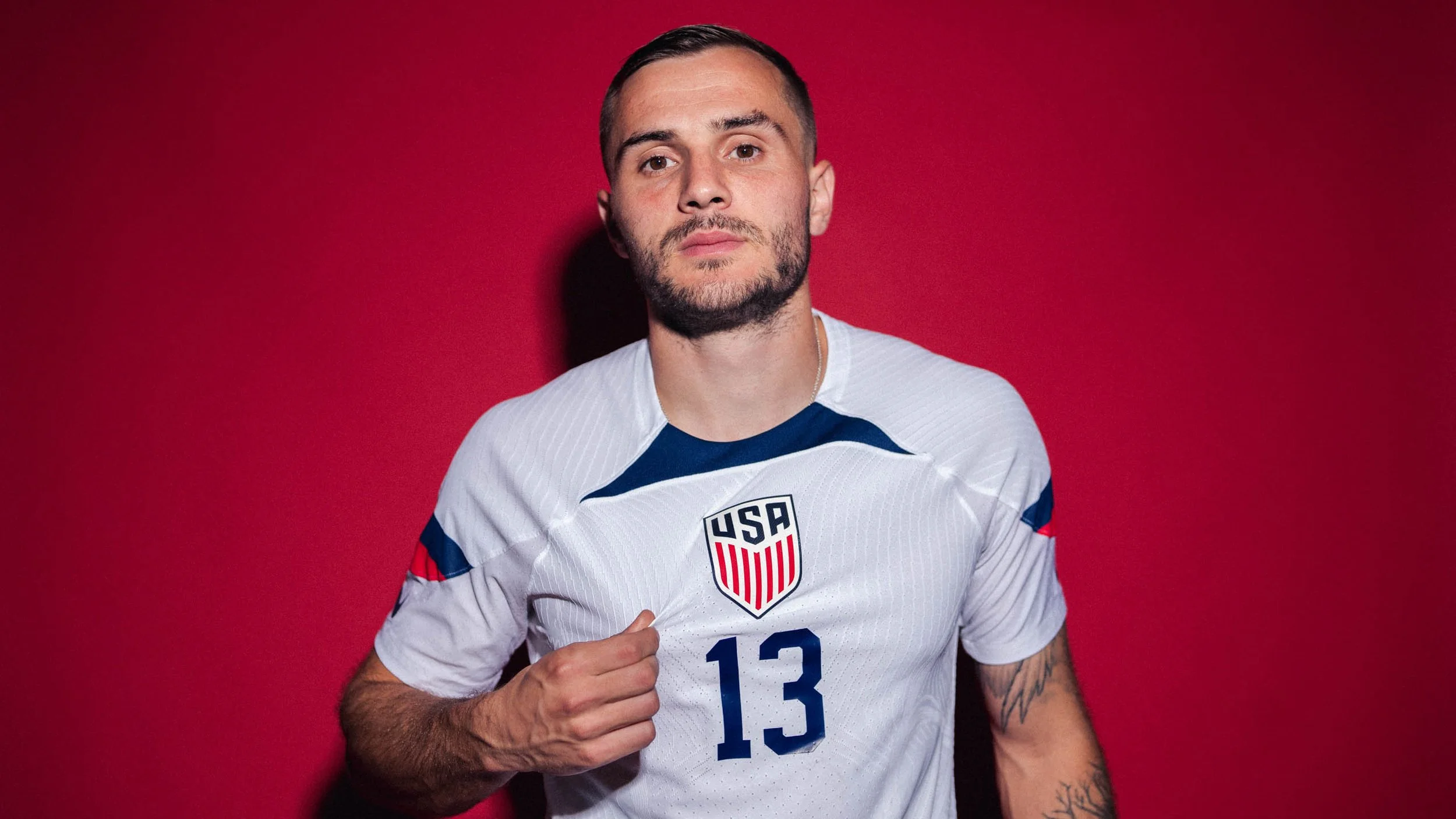 A man in a USA soccer jersey against a red background, posing with a fist.