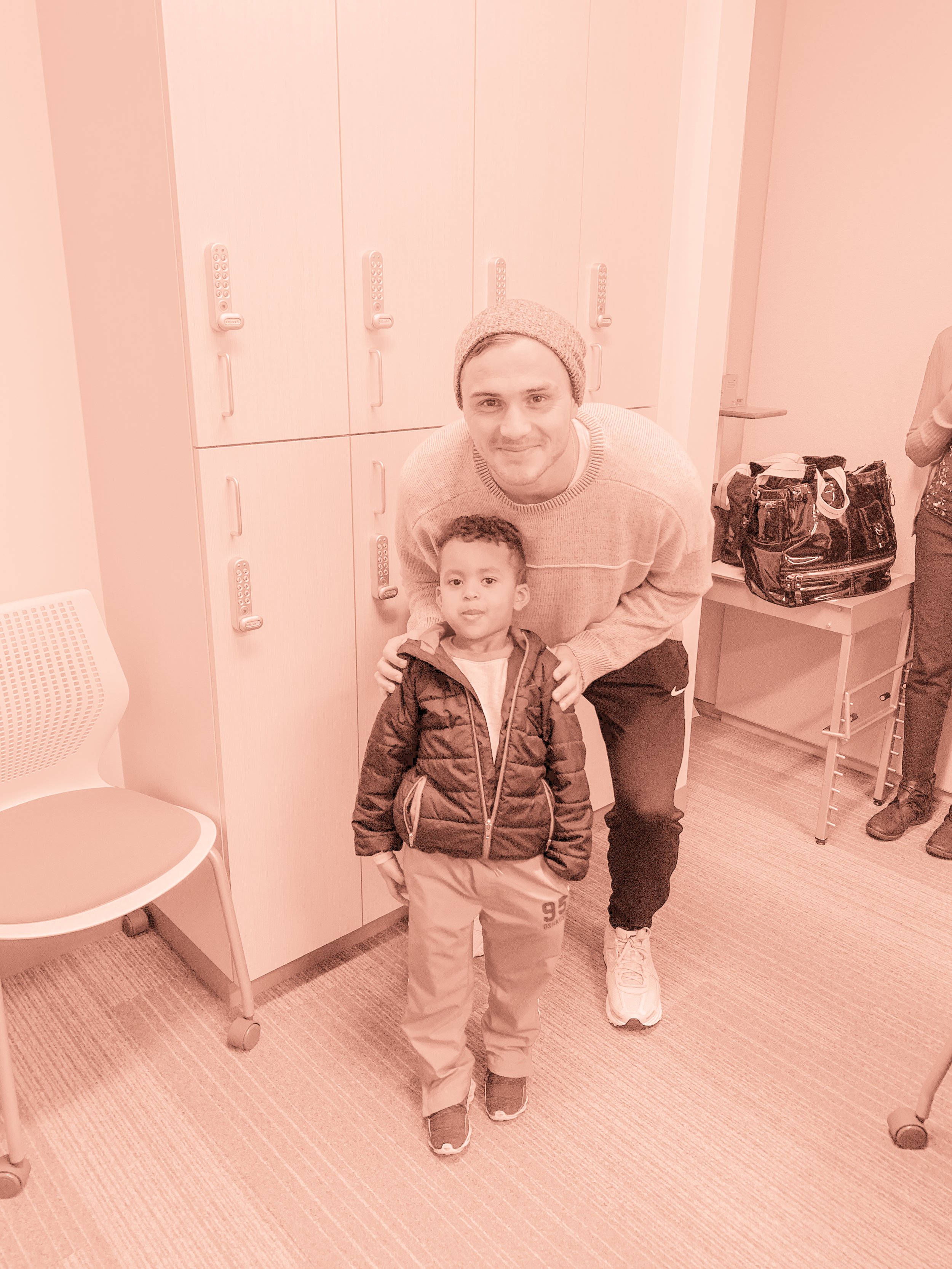 A man and a young boy standing indoors, in front of lockers, with a chair to their left, and a black bag on a small table to their right.