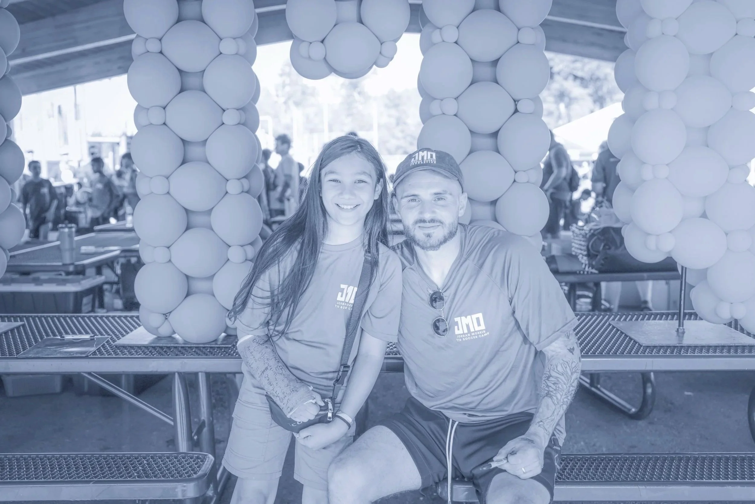 A young girl and a man sitting together, smiling, at a table with balloon decorations around them. The scene is bright and cheerful, likely at an outdoor event or celebration.