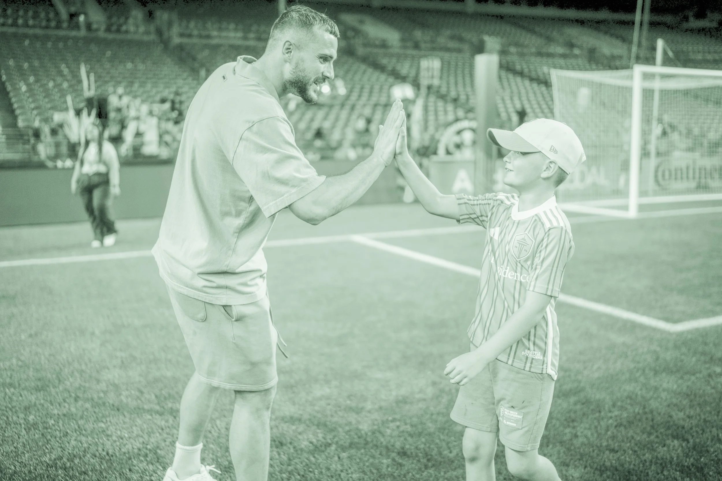 A man and a young boy are giving each other a high five on a sports field. The man has short hair and a beard, wearing a light-colored t-shirt and shorts. The boy is wearing glasses, a baseball cap, and a striped sports jersey. In the background, there are empty stadium seats and a goalpost.
