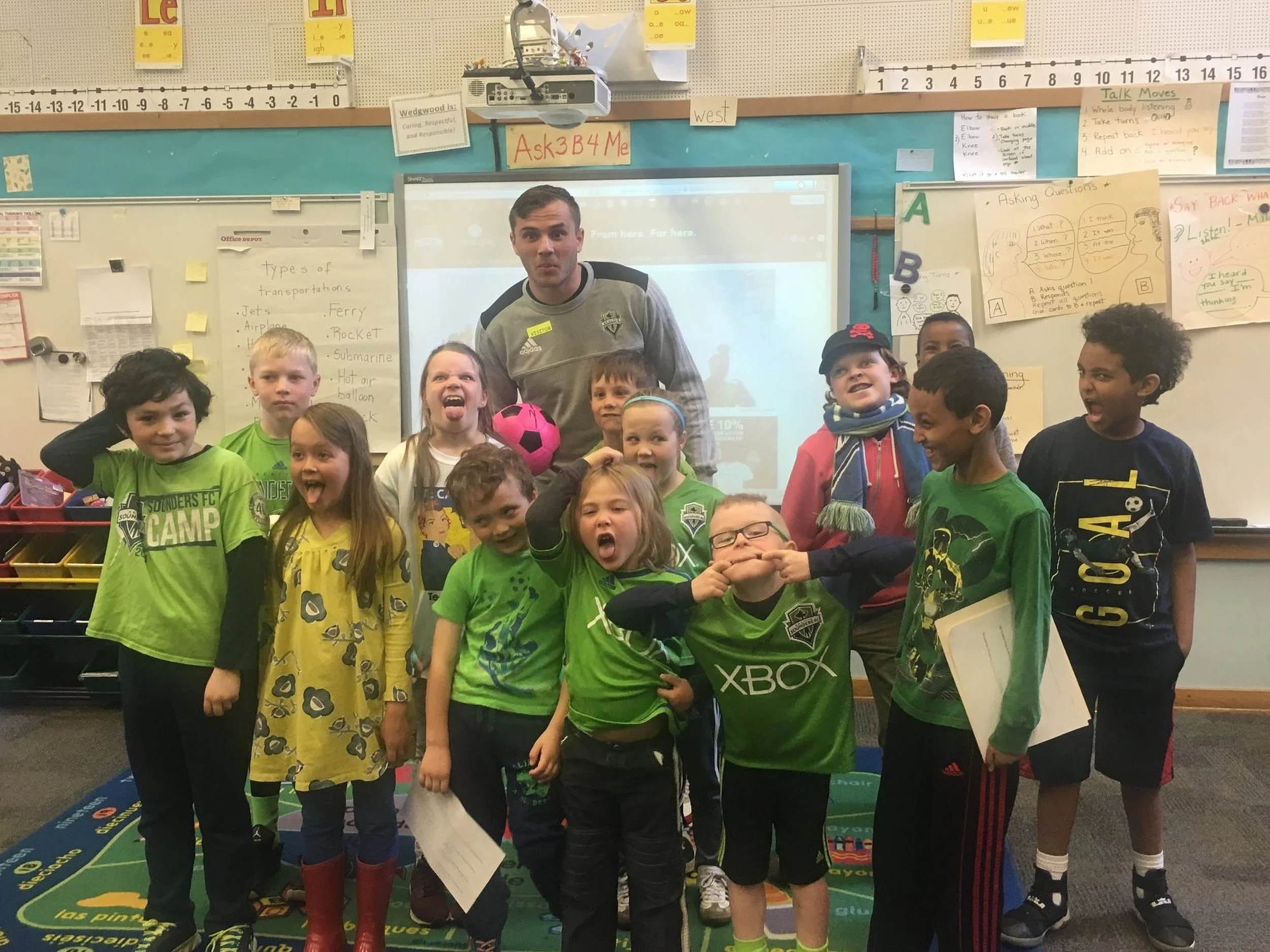 A group of children standing in a classroom, some wearing green sports shirts and others in casual clothing, with a man in a gray jacket standing behind them. The classroom has educational posters, a whiteboard, and a projector overhead.