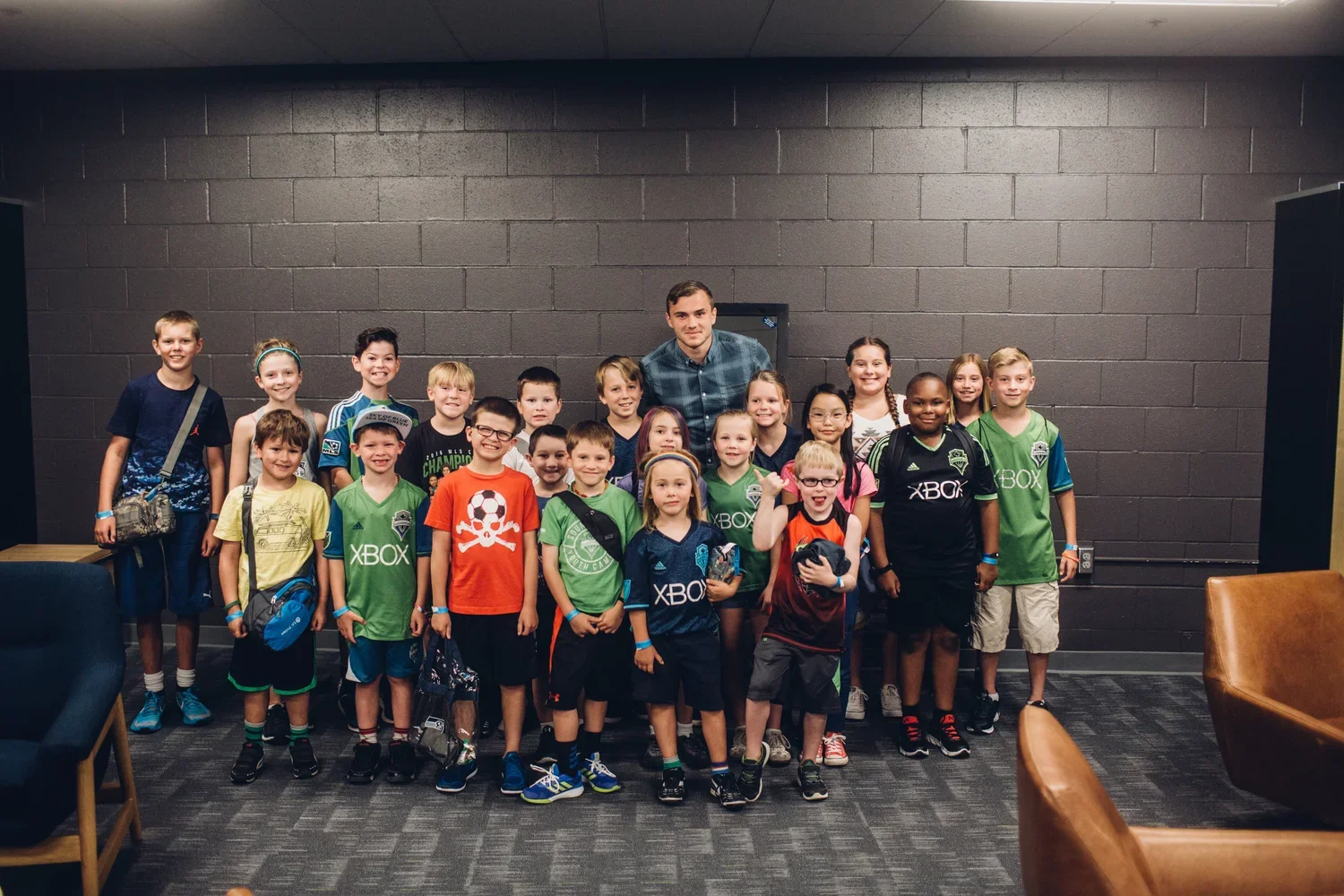 Group of children and a young adult, likely a coach or teacher, posing indoors against a dark gray brick wall. The children are smiling, some wearing sports jerseys, and a few are holding backpacks.