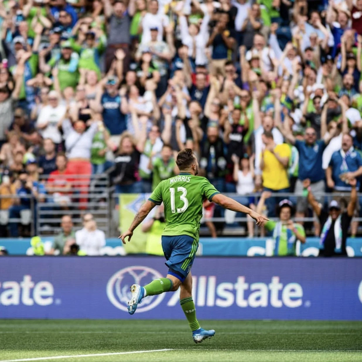 Soccer player wearing a green jersey with the name 'MORRIS' and the number 13 dribbling a ball on the field during a game with a large crowd of spectators in the stands.