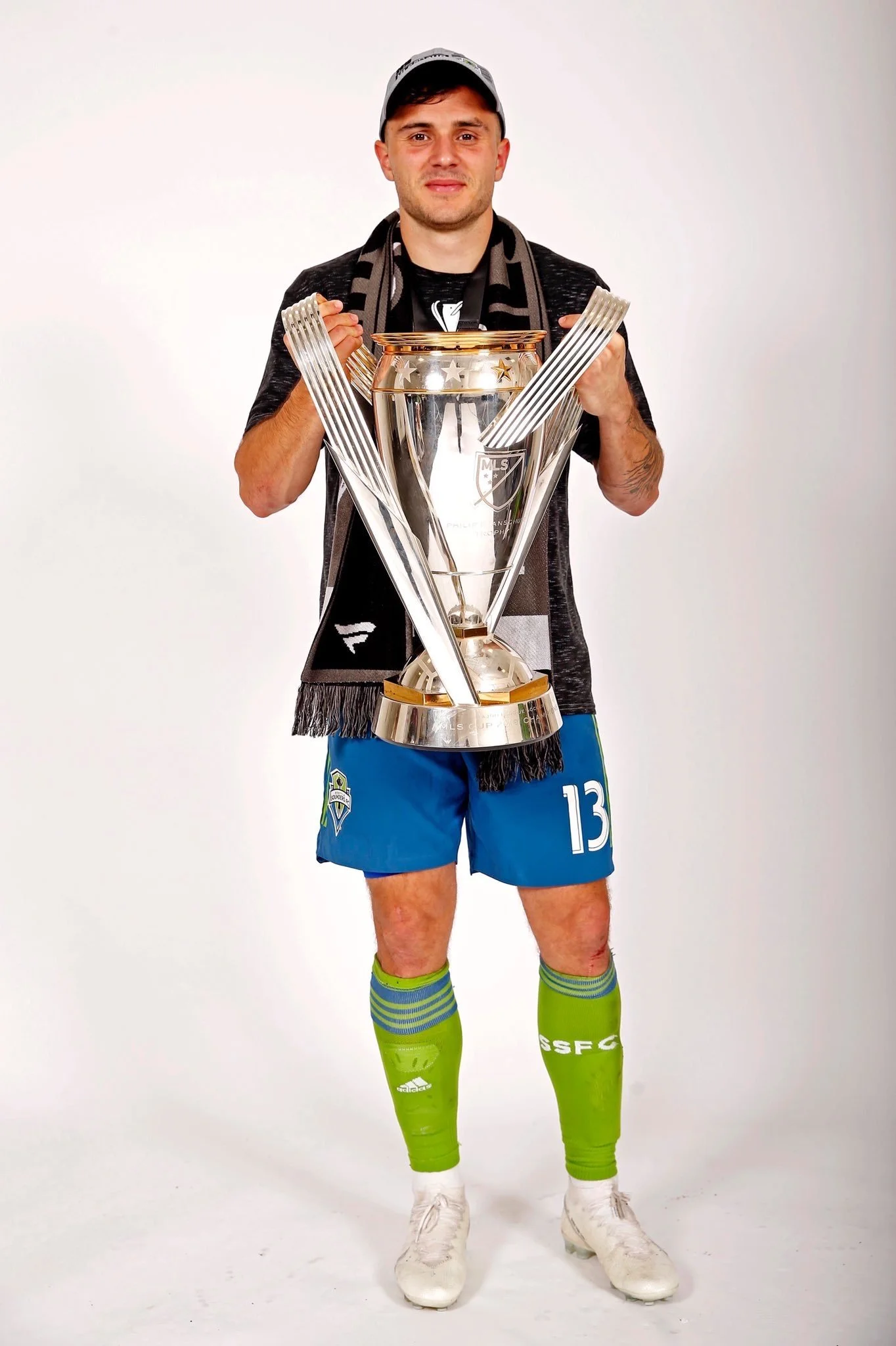 A man in a soccer uniform holding a large trophy, wearing a black sports shirt, blue shorts with a team logo, bright green soccer socks, and white soccer cleats, standing in front of a plain white background.