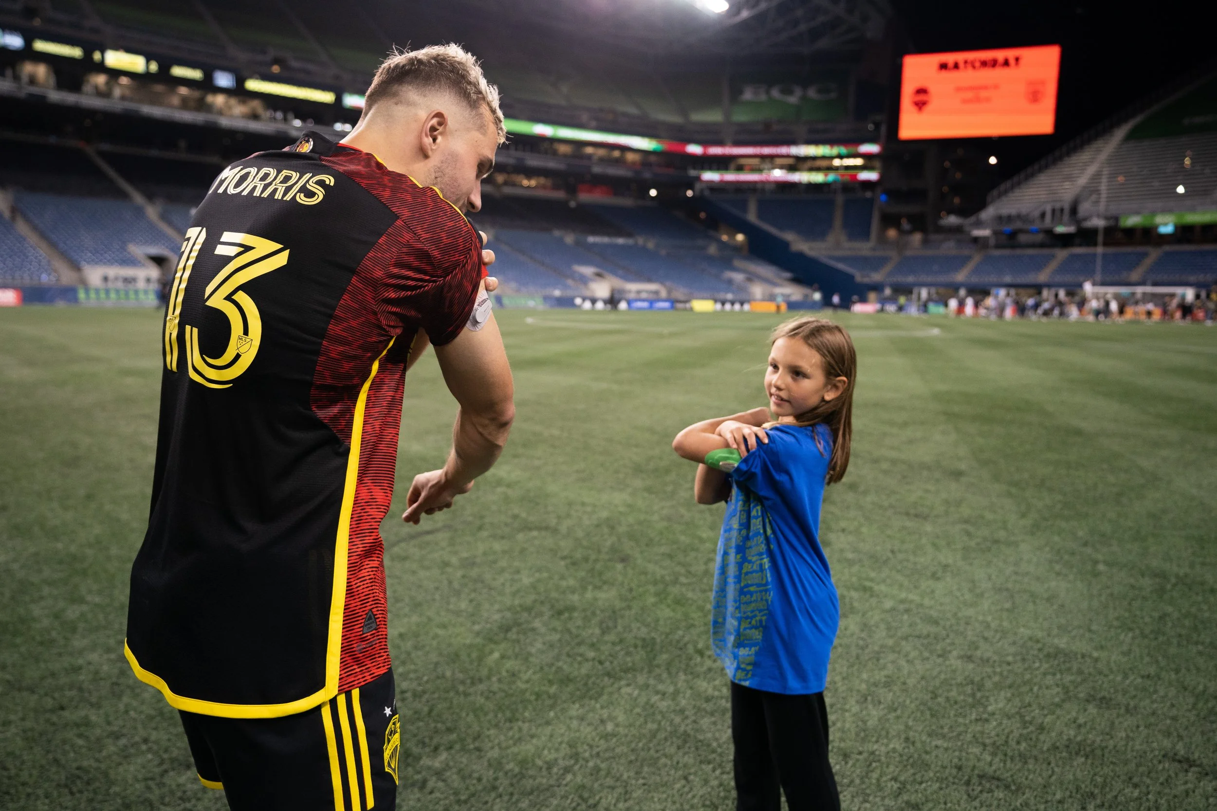 A soccer player wearing a black and red jersey with the name 'MORRIS' and number 13 on it stands on a soccer field, speaking to a young girl wearing a blue shirt, inside a large stadium at night.
