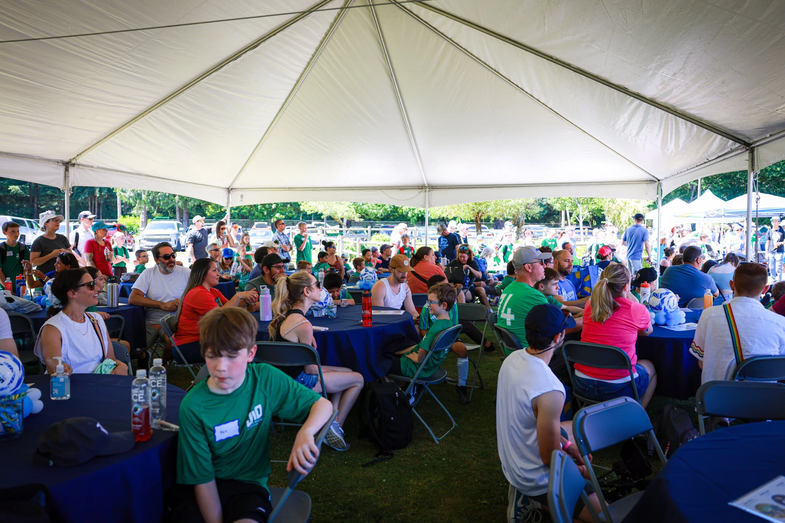 A large outdoor gathering under a white canopy tent with people sitting at tables and others standing around, likely at a community event or festival, with trees and parked cars in the background.