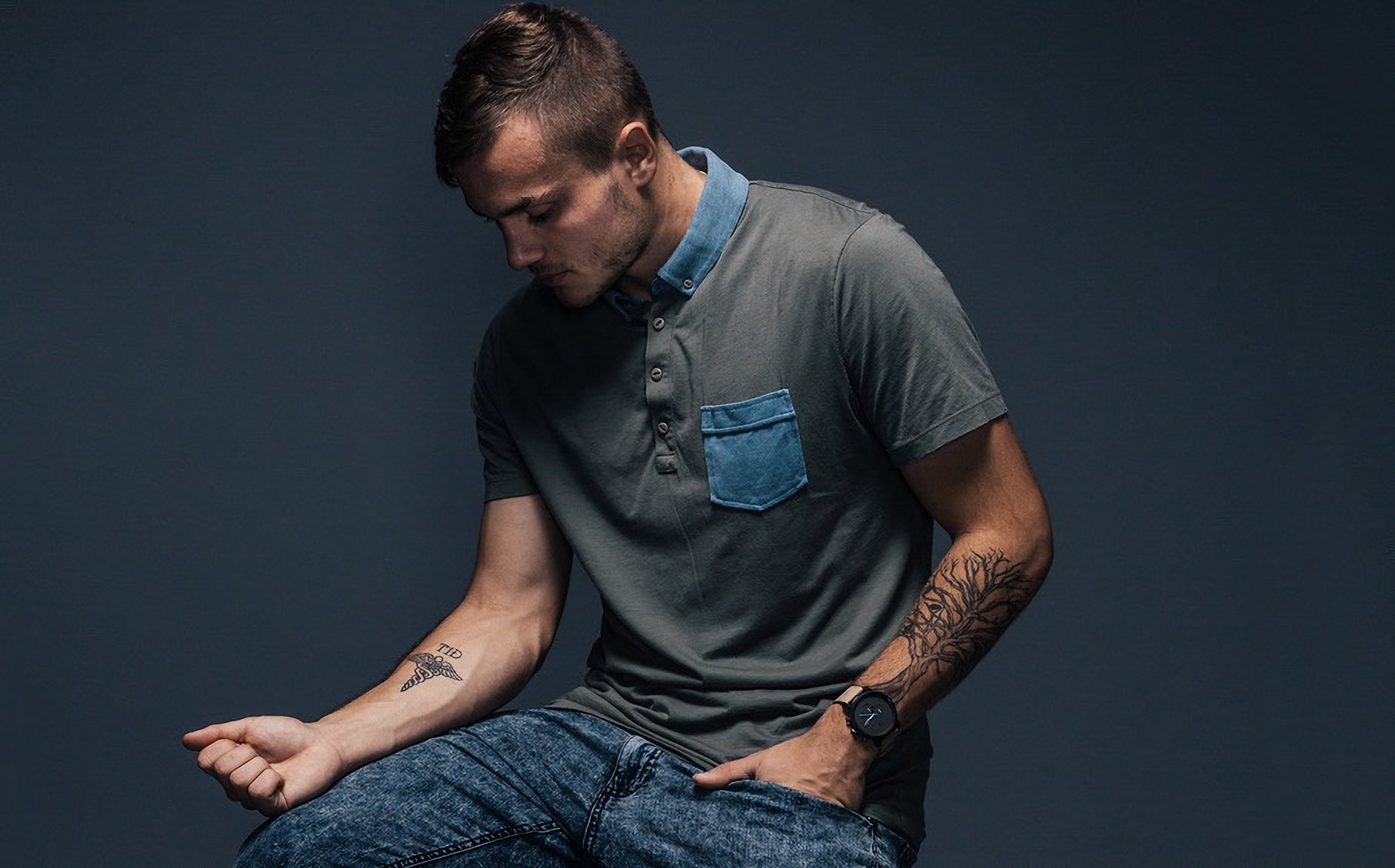 A young man with tattoos on his arms and a short hairstyle, wearing a grey shirt with a blue collar and a blue pocket, sitting against a dark background.